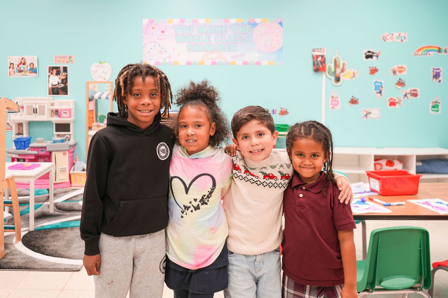 Four diverse elementary school children smiling together in a colorful classroom setting.