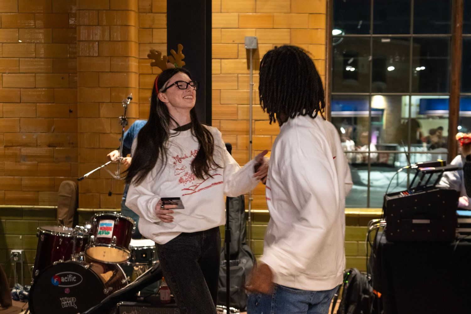 Two friends chat and laugh near a drum set in a warmly lit brick-walled venue during a live music event.
