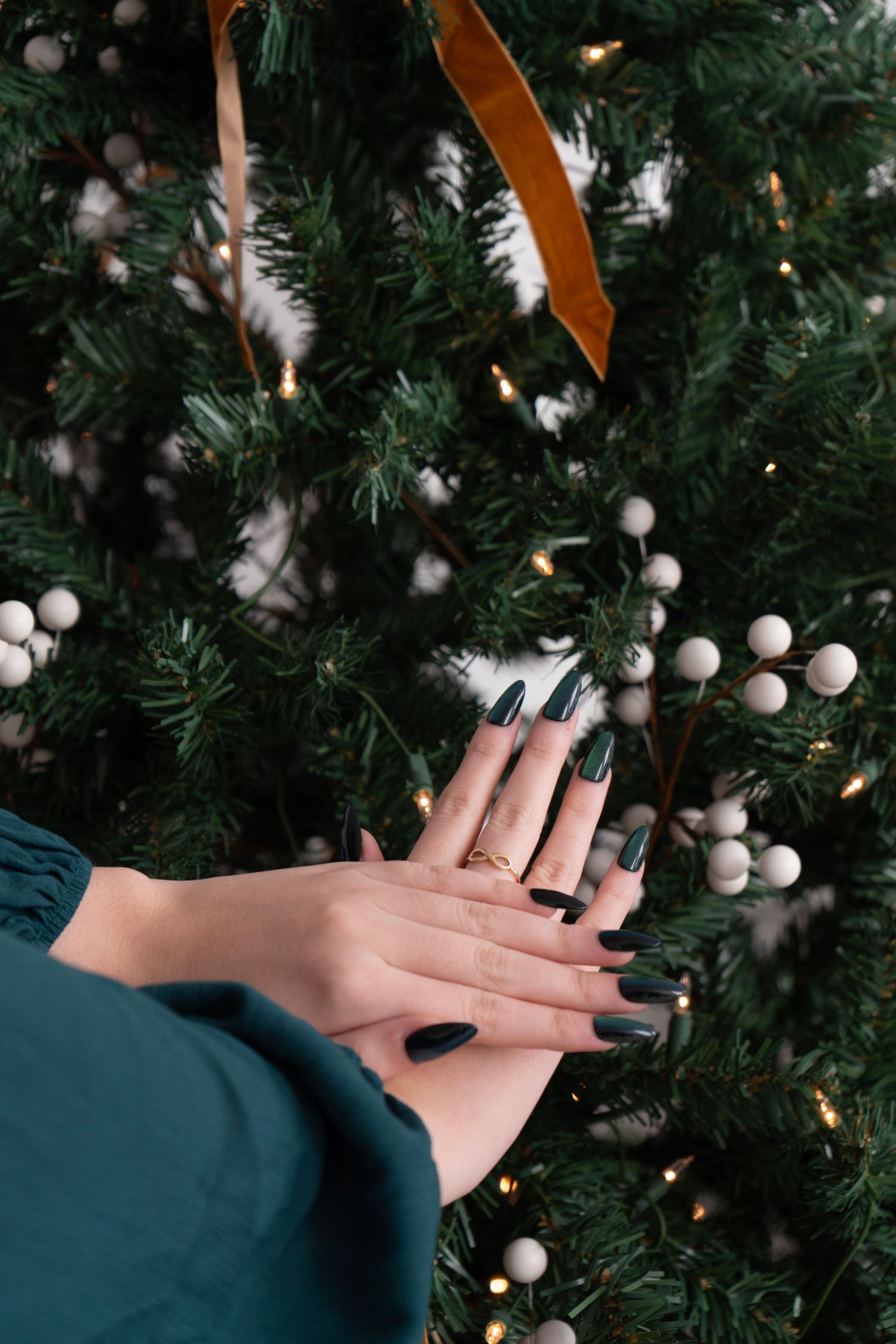 Woman with dark green manicure and gold ring poses hands in front of decorated Christmas tree with white berries.
