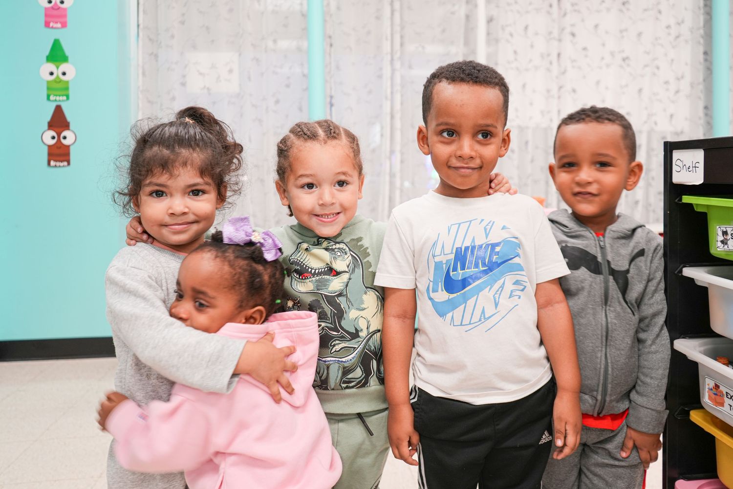 Five young children smiling and posing together in a colorful classroom setting.