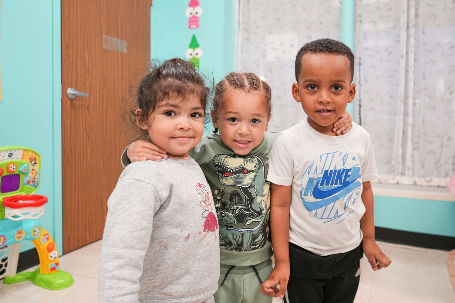 Three young children smile and pose together in a colorful classroom setting, one wearing a Nike shirt.