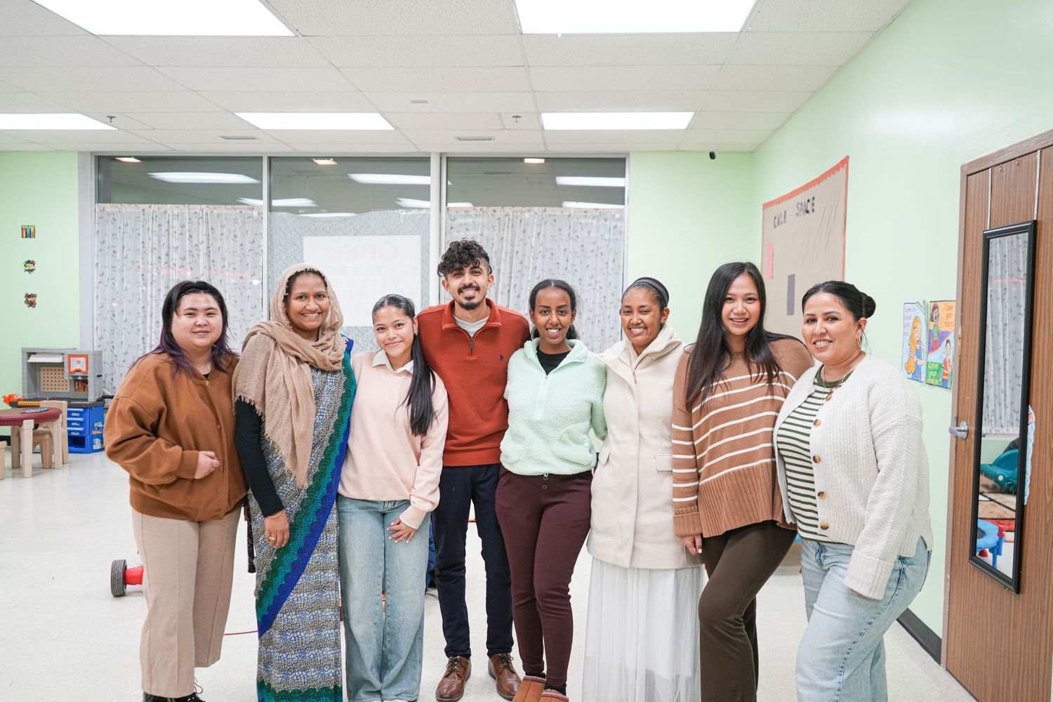 Diverse group of eight smiling people posing together in a bright classroom setting.