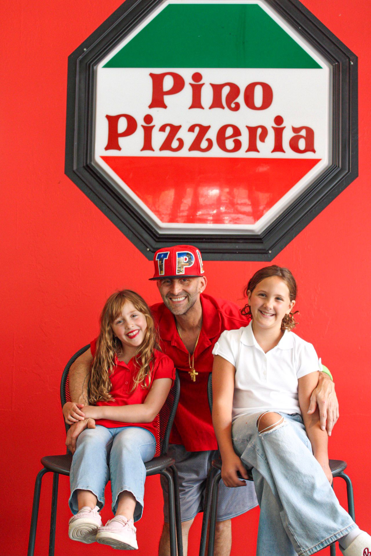 A man and two girls smile together in front of a red Pino Pizzeria sign.