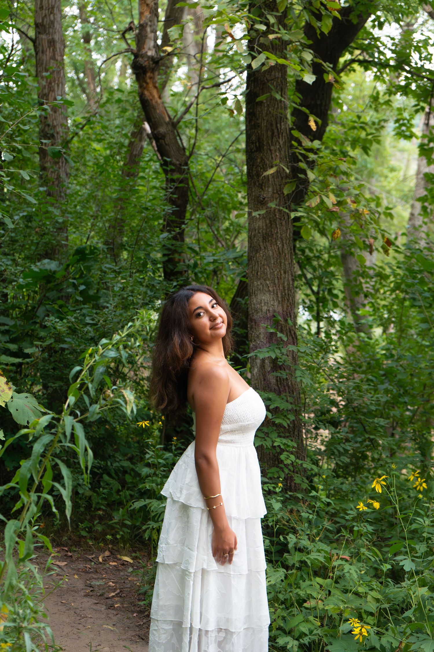 Young woman in white tiered dress posing gracefully on a forest path surrounded by lush green trees.