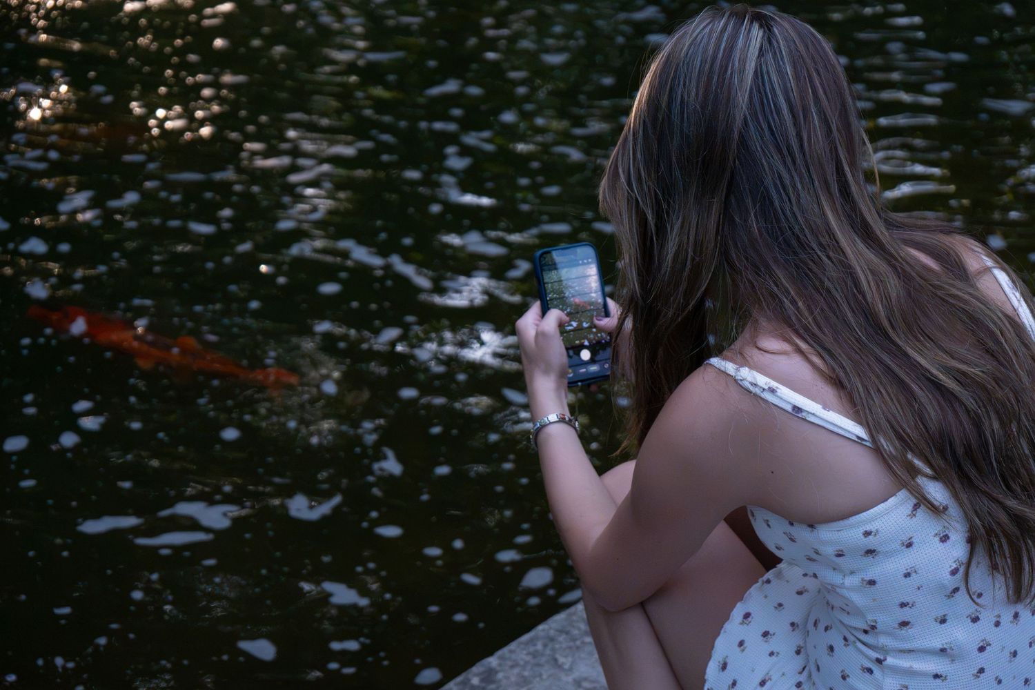 A young woman photographs colorful koi fish in a pond using her smartphone while sitting at the water's edge.