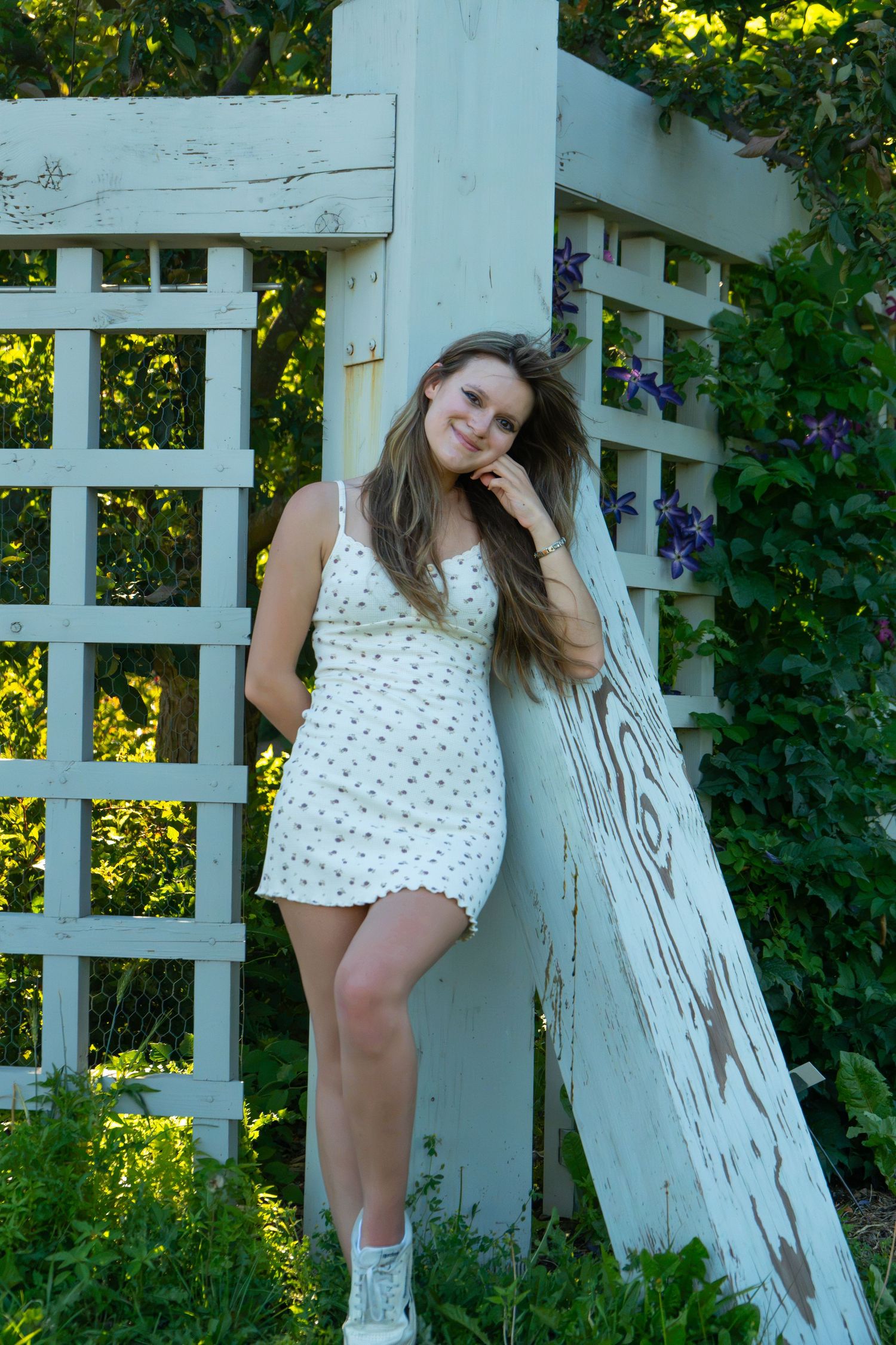Young woman in white floral sundress leaning against rustic garden trellis surrounded by lush greenery and purple flowers.
