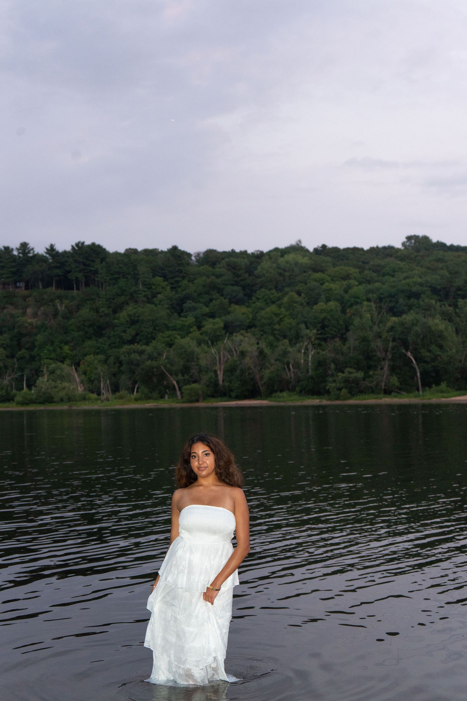 Young woman in white strapless dress standing in calm lake water with lush green forest backdrop.
