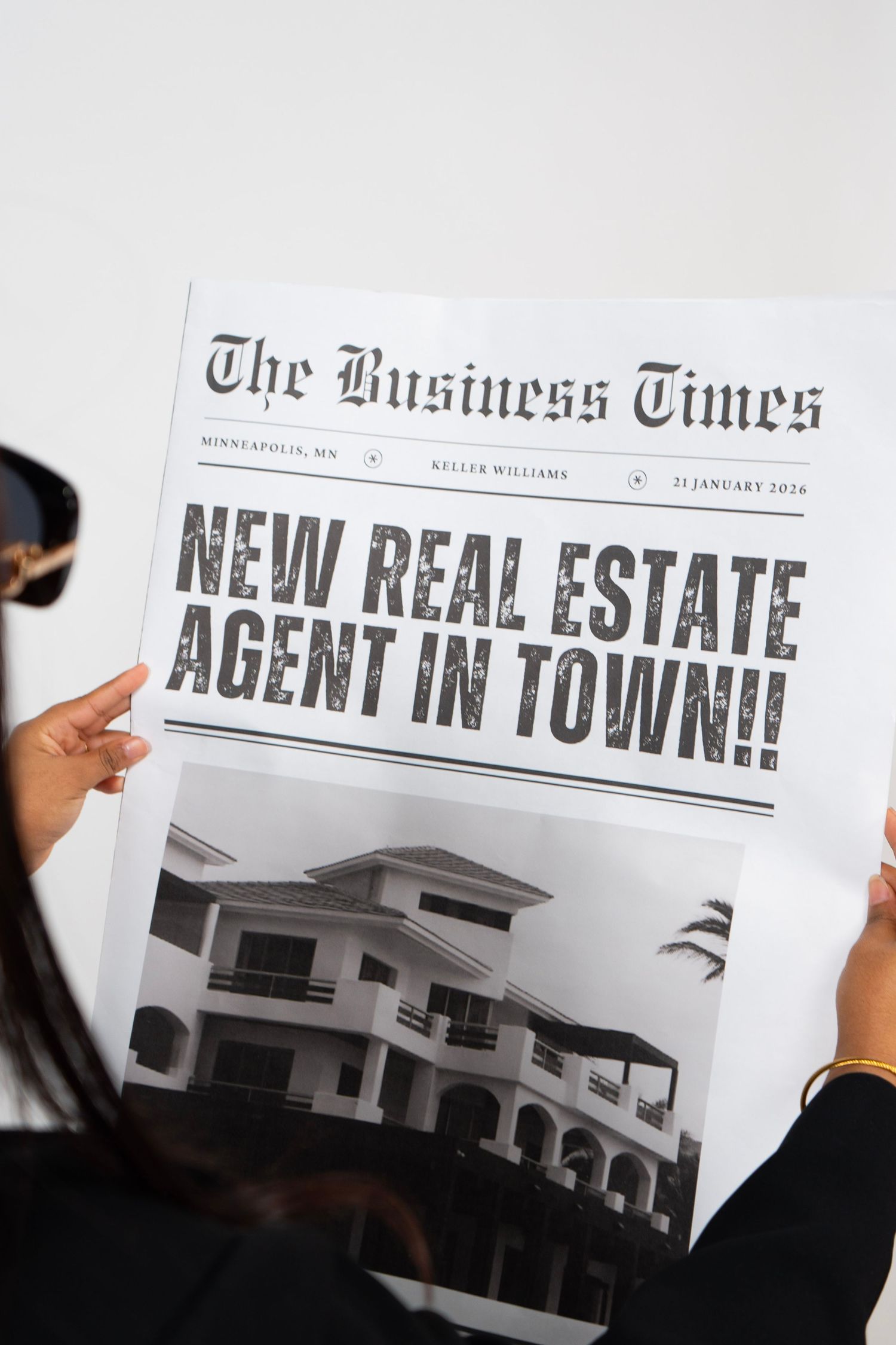Person holding The Business Times newspaper with headline NEW REAL ESTATE AGENT IN TOWN featuring a house photo.