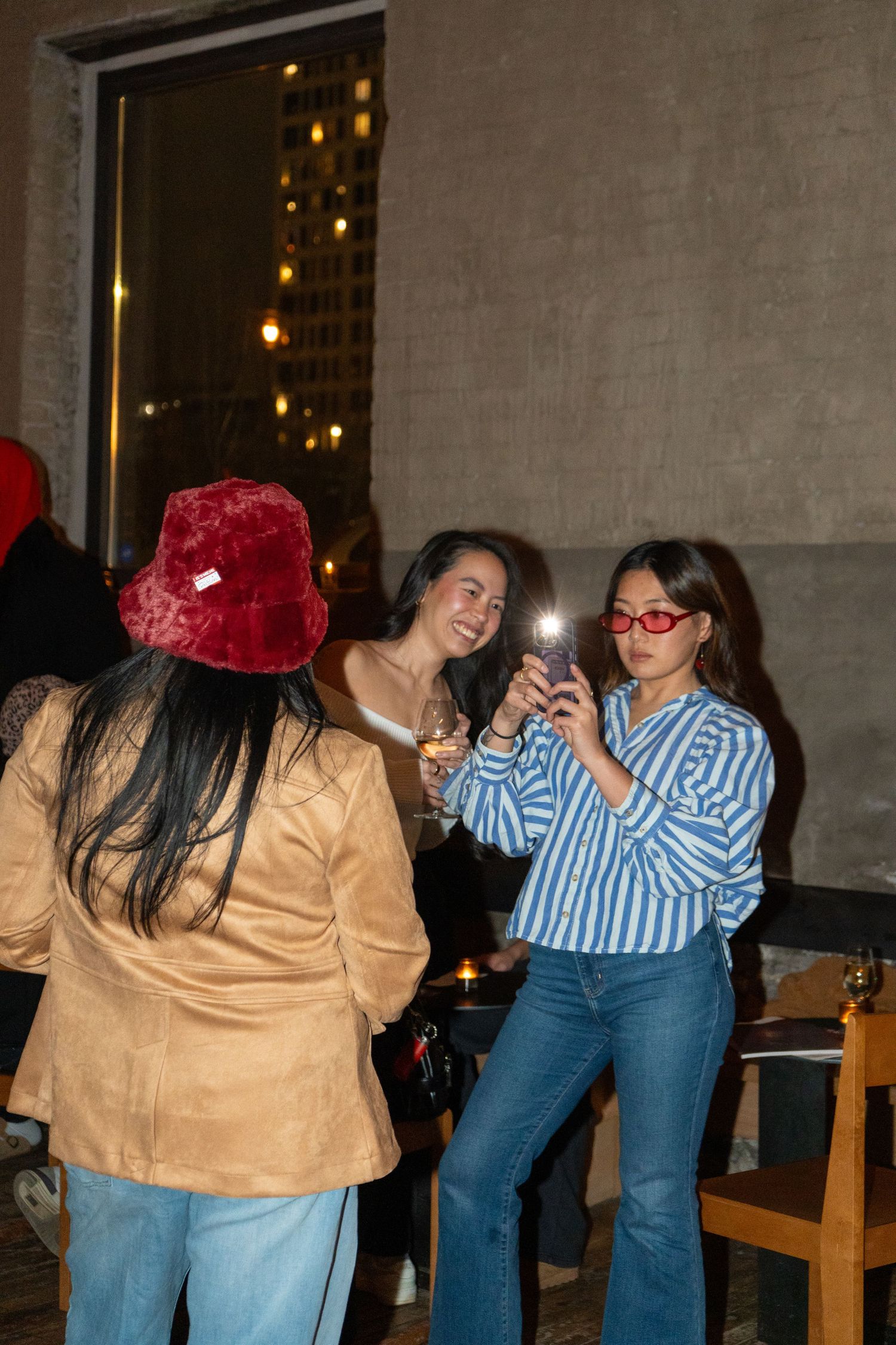 Three young women socializing at a rooftop bar, one taking photos with smartphone flash, wearing trendy outfits and red accessories.