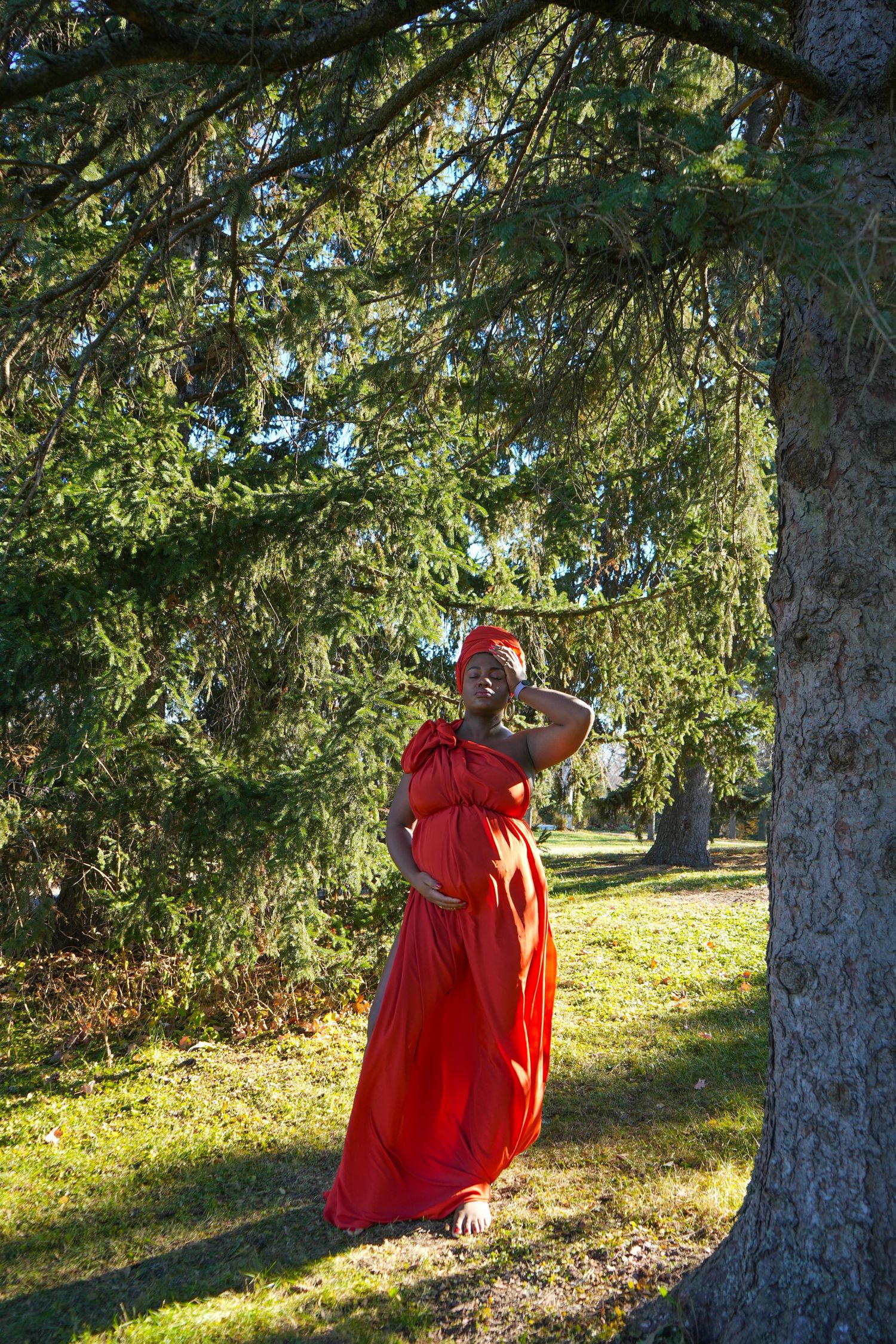 Pregnant woman in red dress and headwrap stands under pine tree in sunny park, shielding eyes from light.