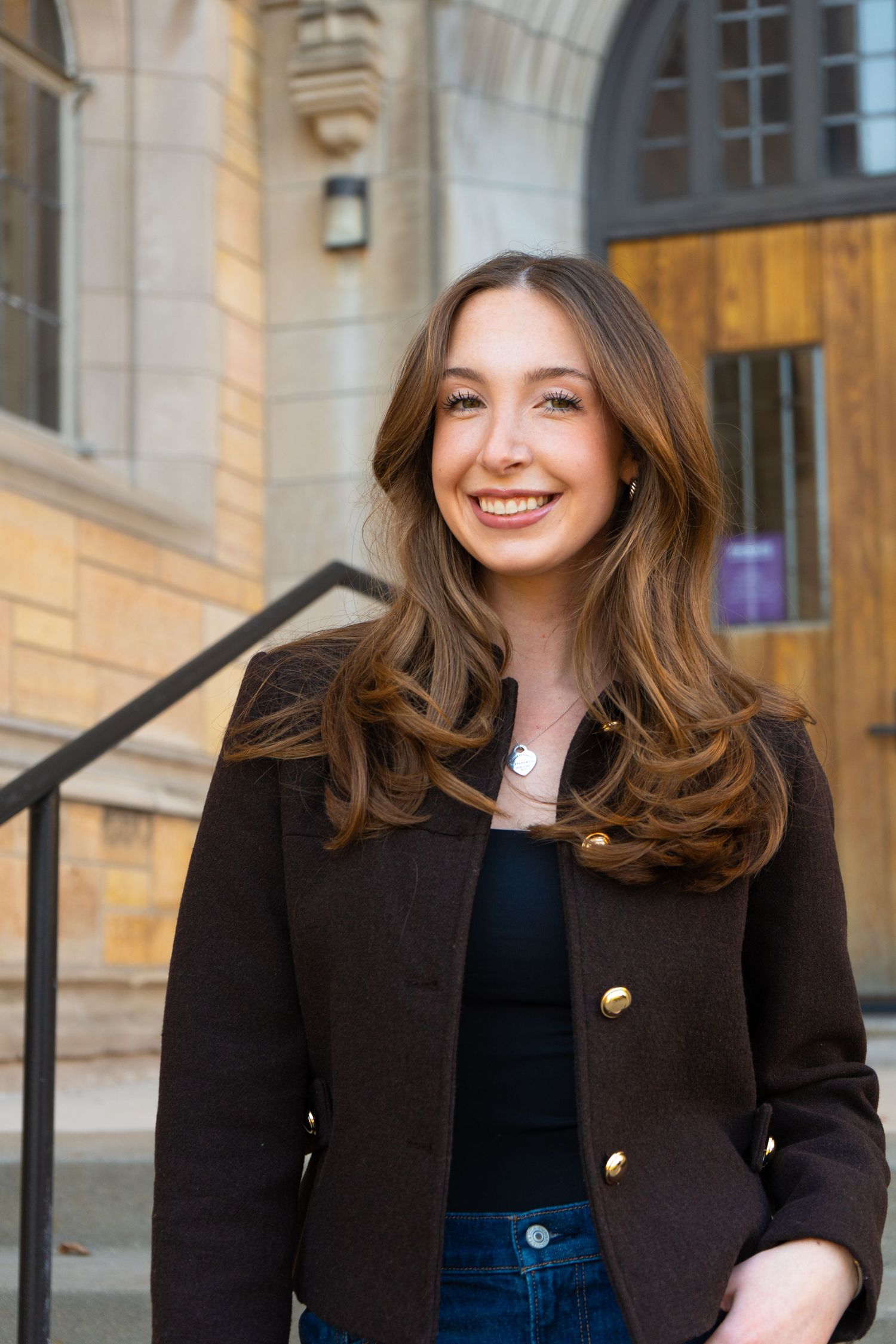 Smiling young woman with long brown hair wearing a dark blazer standing outside a stone building.