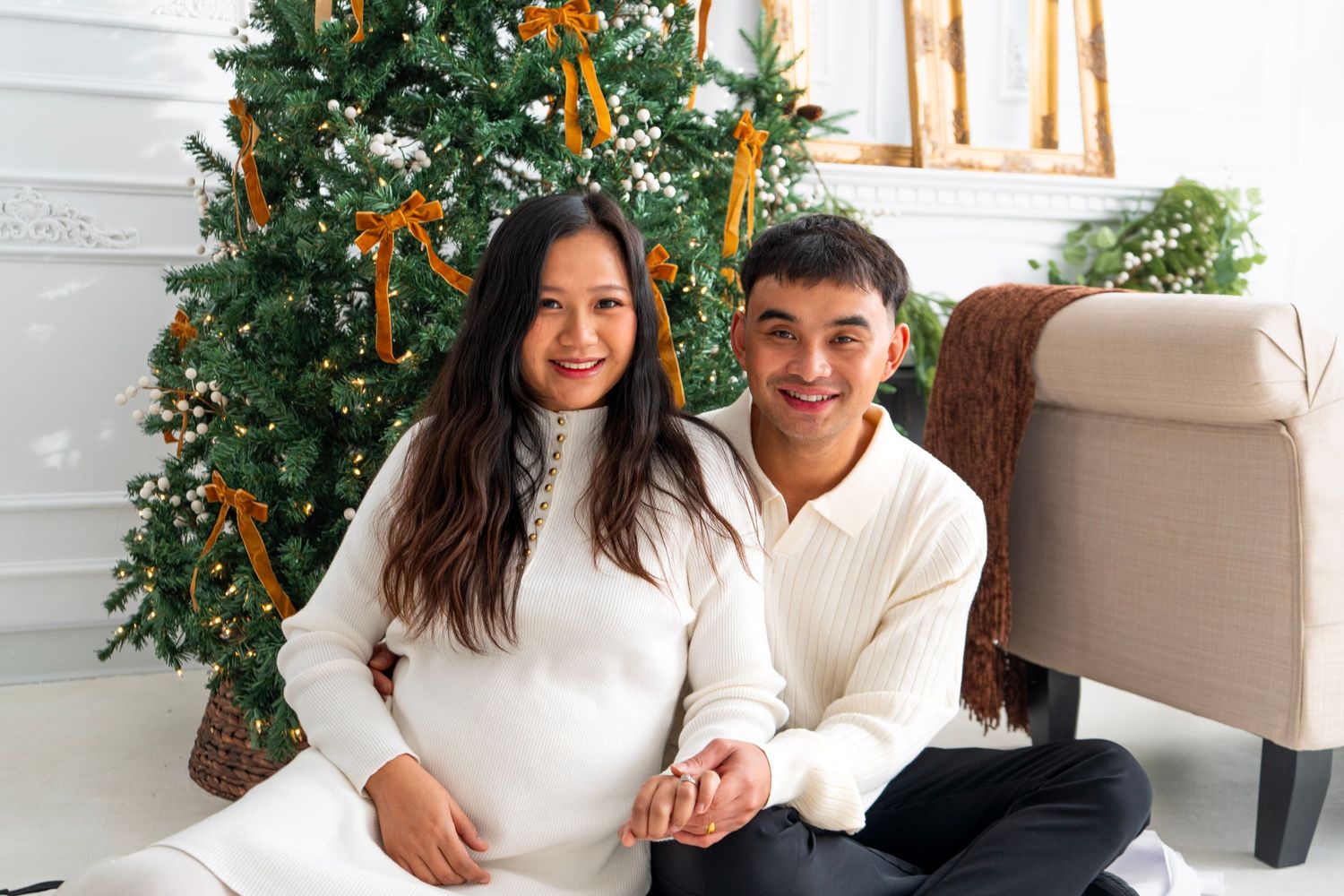 Smiling couple in white sweaters sitting together in front of a decorated Christmas tree with golden ribbons.