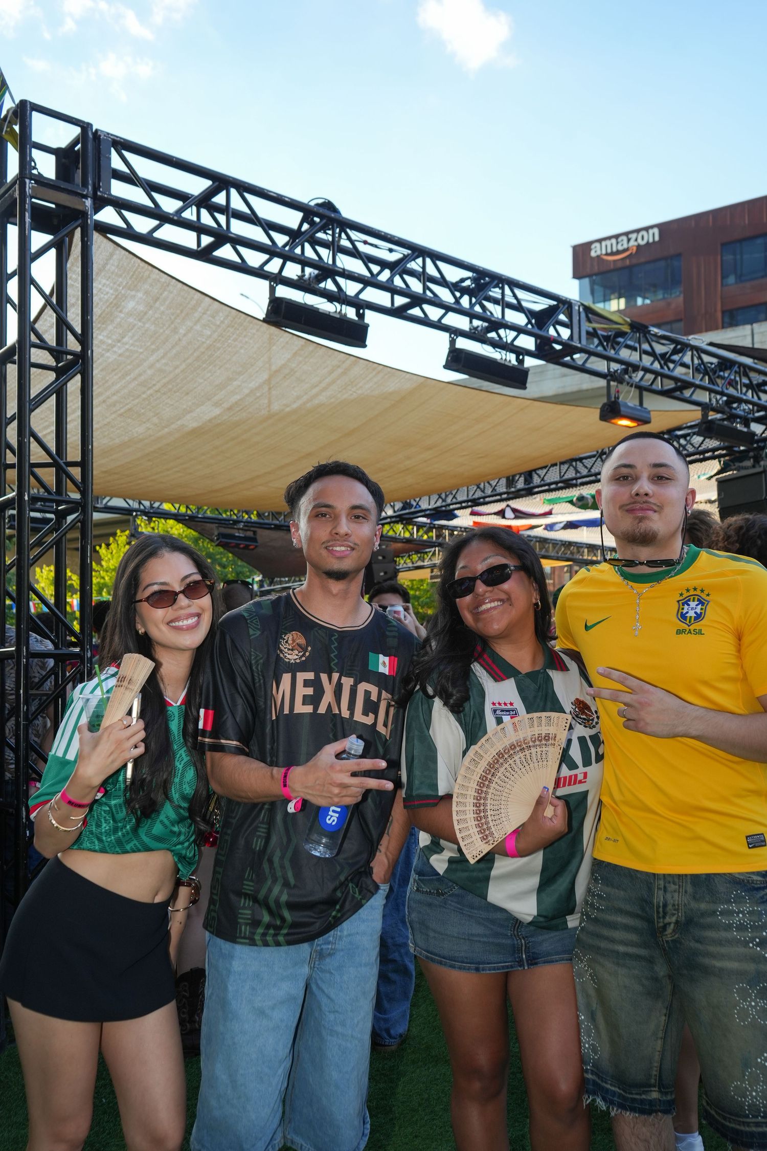 Four friends pose outdoors wearing Mexico and Brazil soccer jerseys at an Amazon-sponsored sports event.
