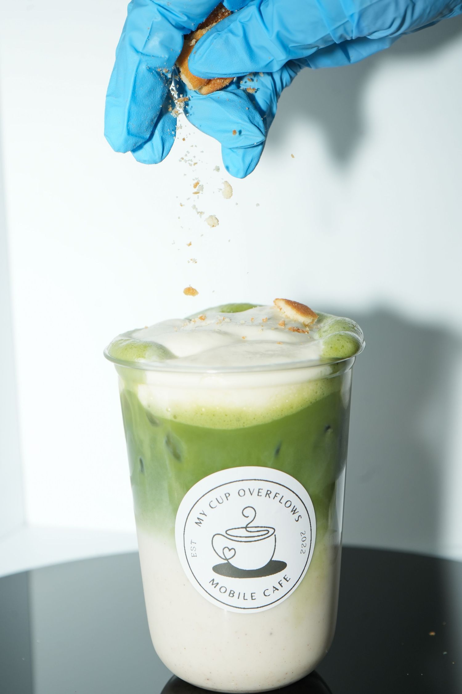 A gloved hand crumbles a cookie over a creamy matcha latte in a branded My Cup Overflows Mobile Cafe cup.