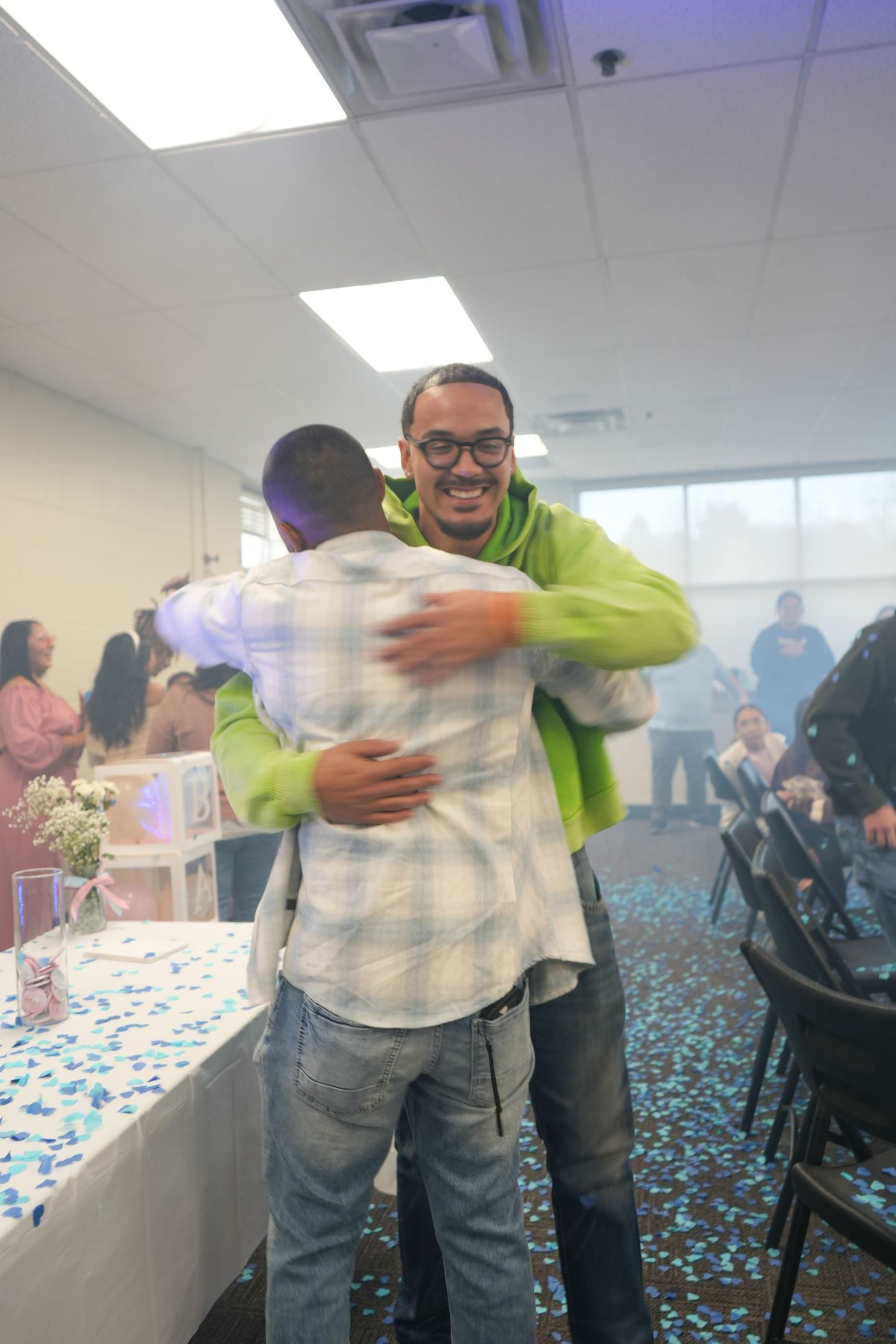 Two men hugging at a gender reveal party with blue confetti on the floor in a decorated indoor venue.