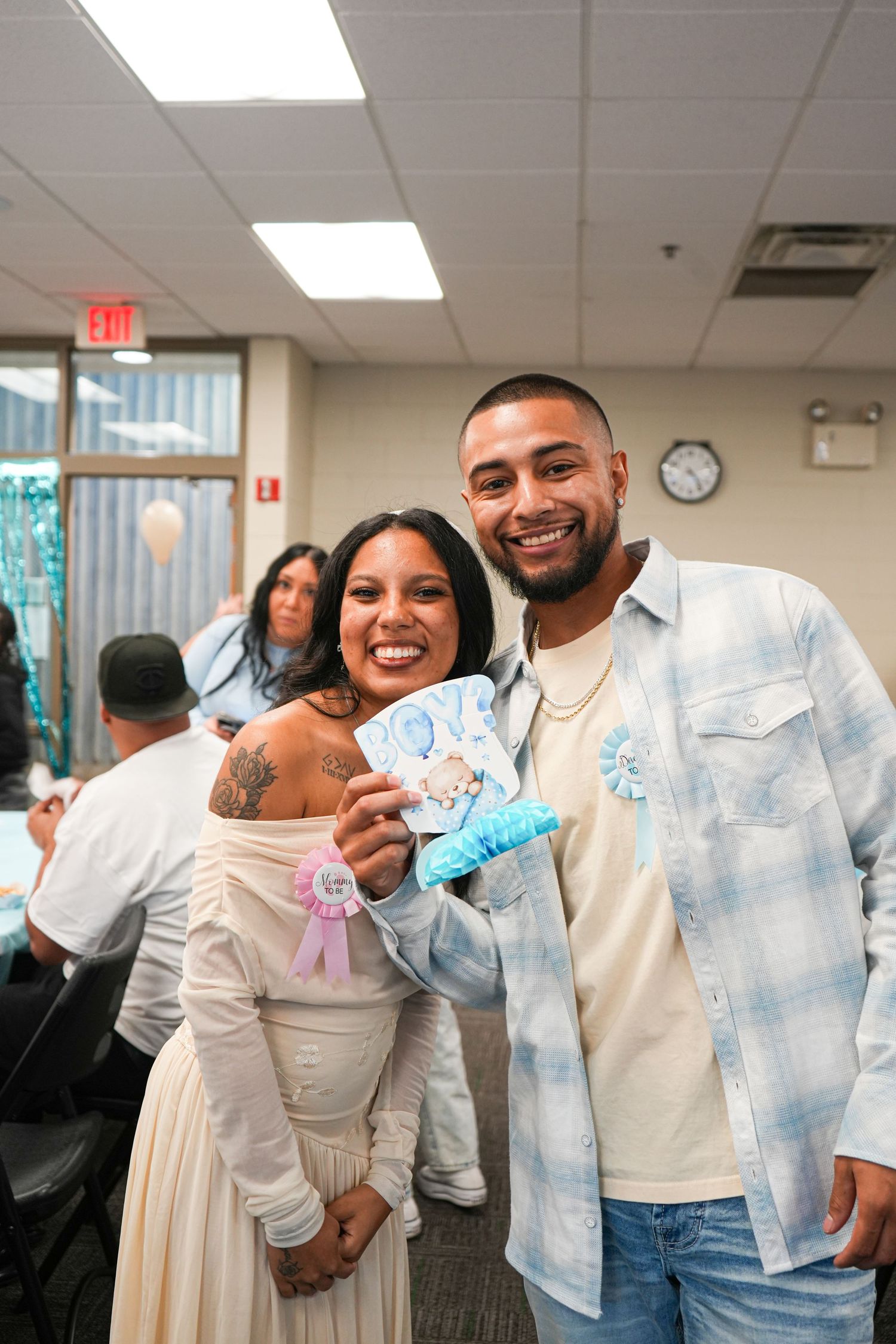 Smiling couple at a baby shower holding blue decorations, woman in white dress with tattoos, man in plaid shirt.