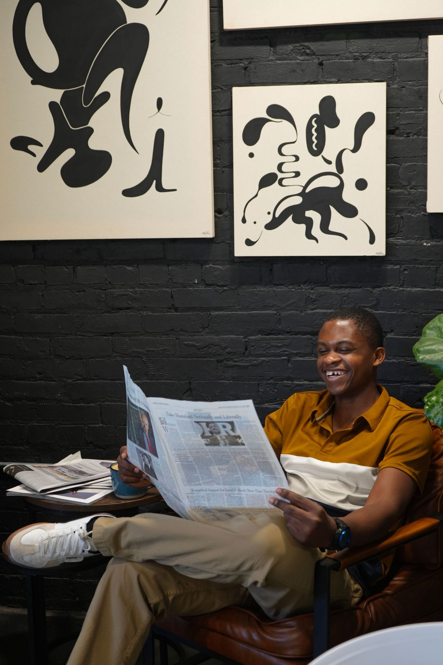 Smiling young Black man reading newspaper in stylish cafe with black brick wall and abstract art prints.