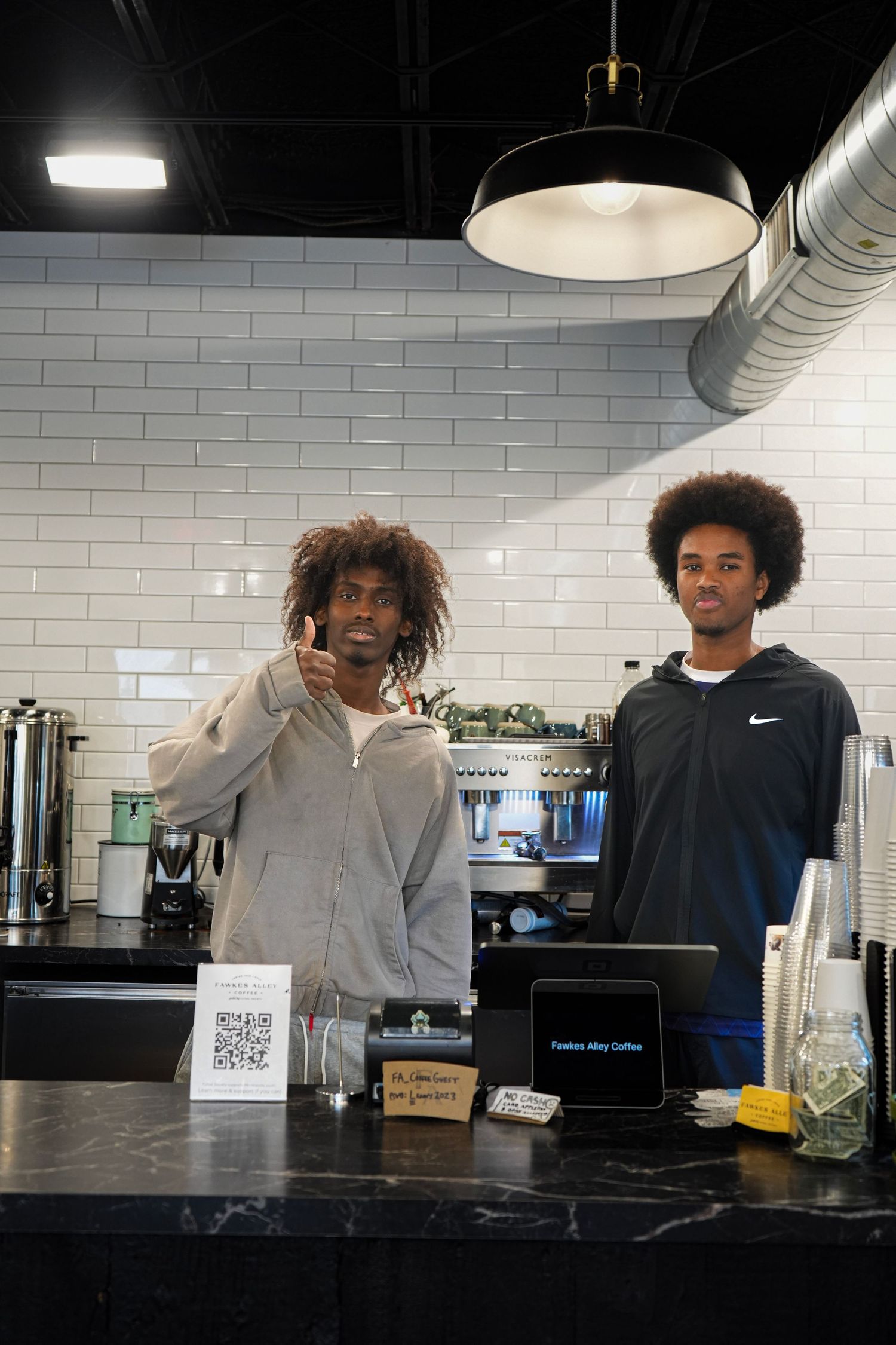 Two baristas stand behind a coffee shop counter with a POS tablet, one giving a thumbs up, white subway tile backdrop.