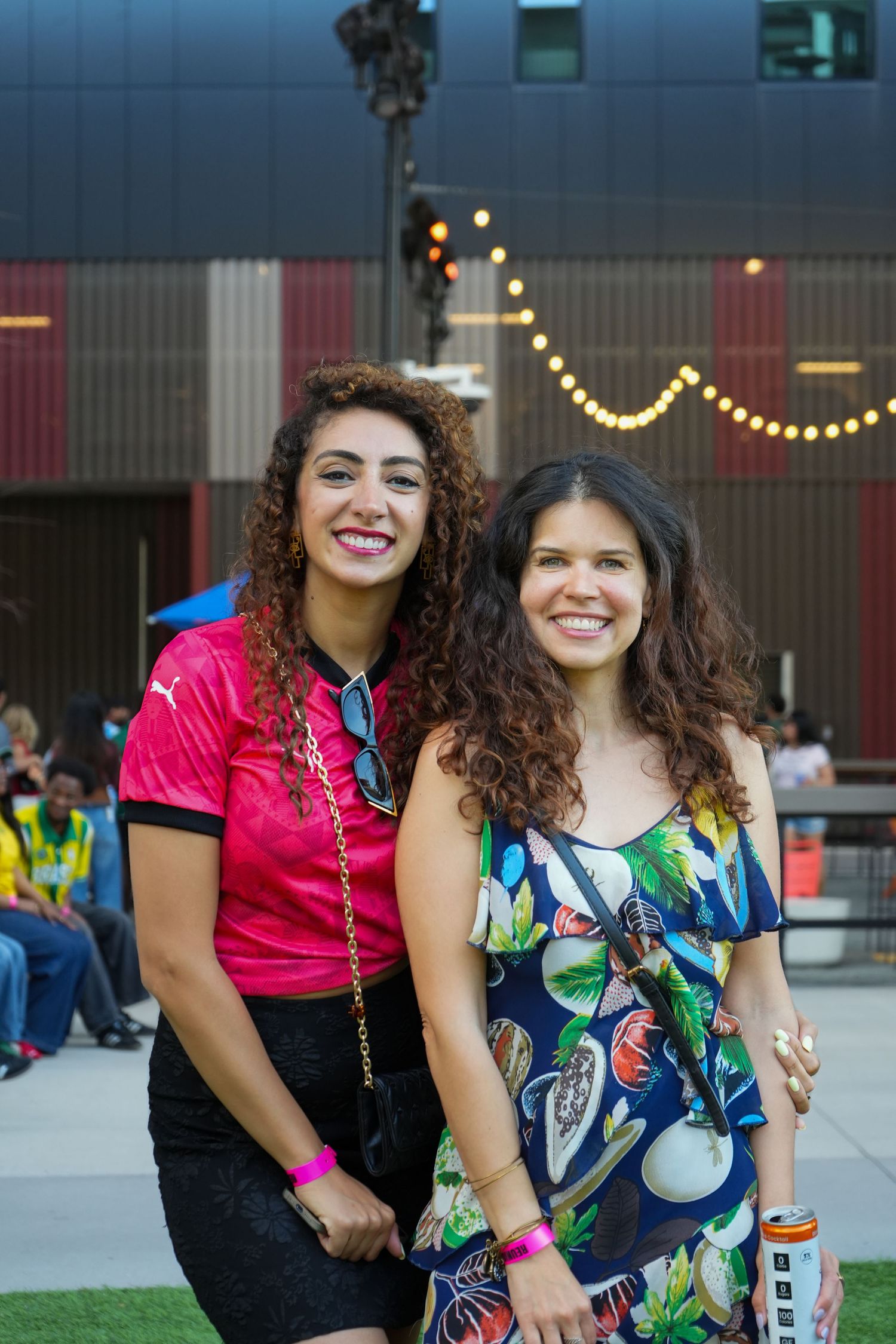 Two smiling women with curly hair pose together outdoors at an evening event with string lights.