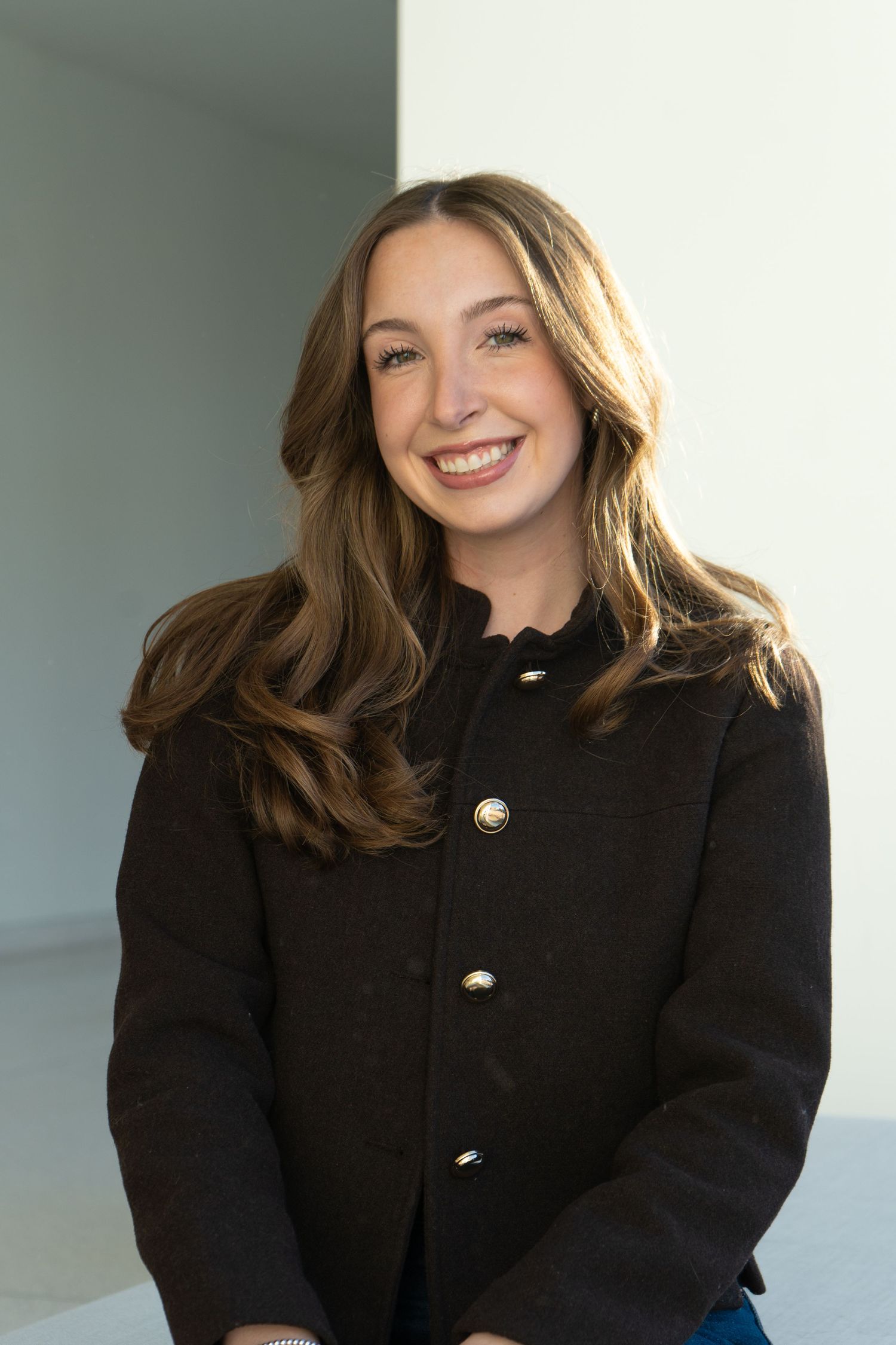 Young woman with long brown hair smiling, wearing a black coat with gold buttons, posed against a light background.