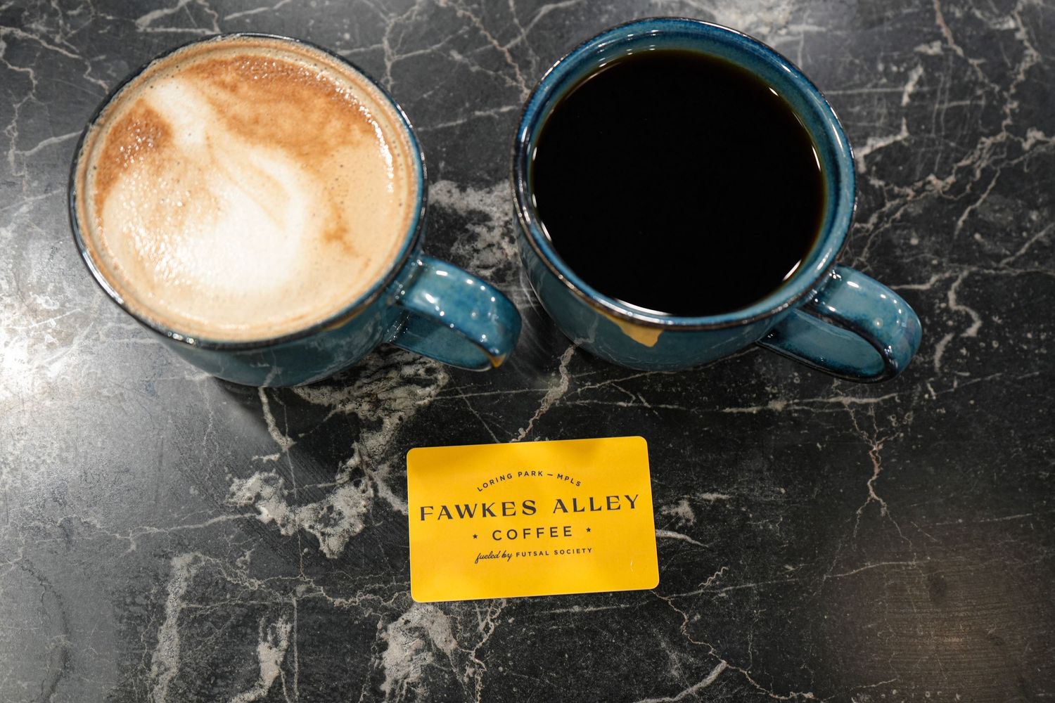 Two blue mugs of coffee beside a Fawkes Alley Coffee gift card on a marble surface.