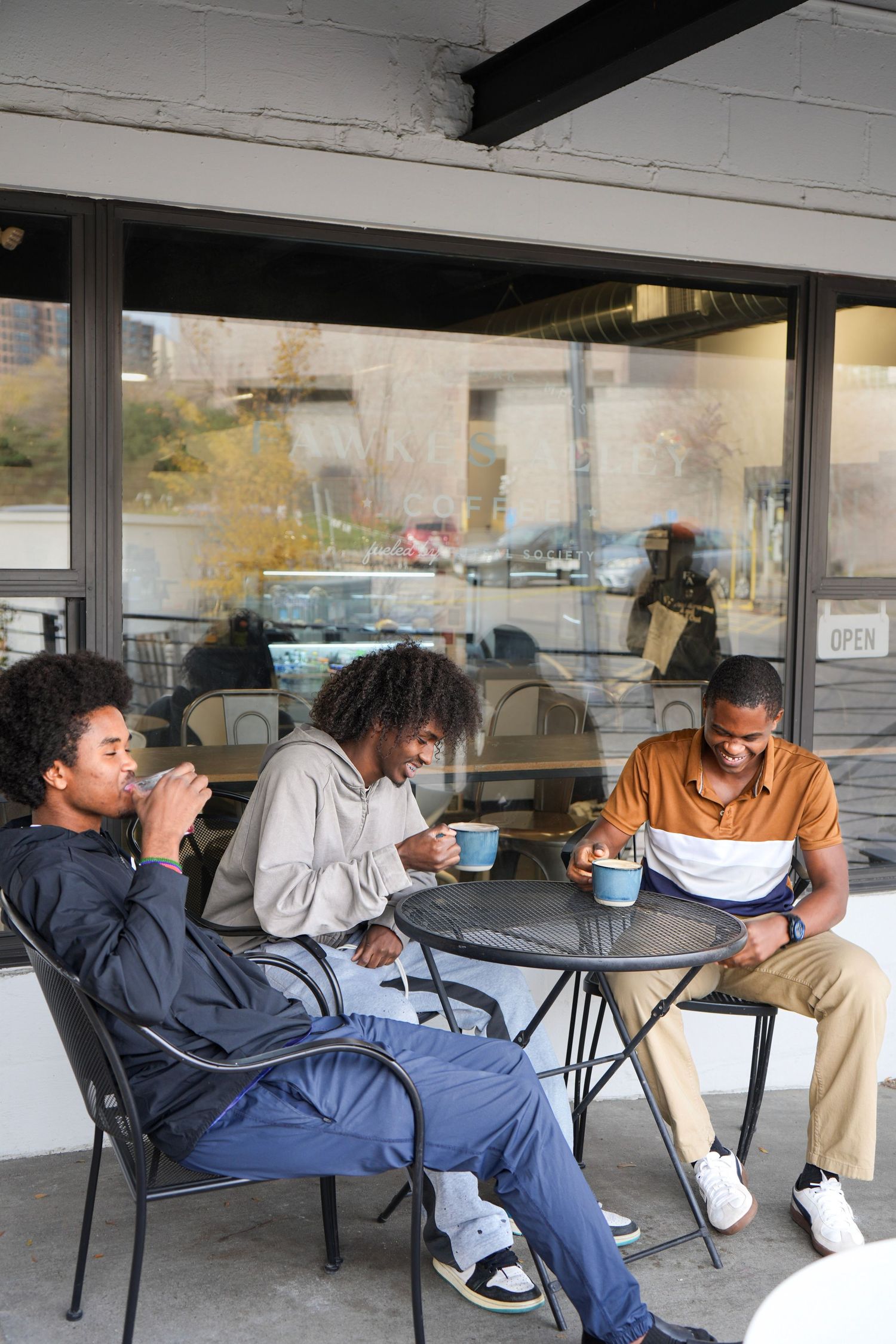 Three young Black men sitting at an outdoor cafe table, enjoying coffee and conversation near a large window.