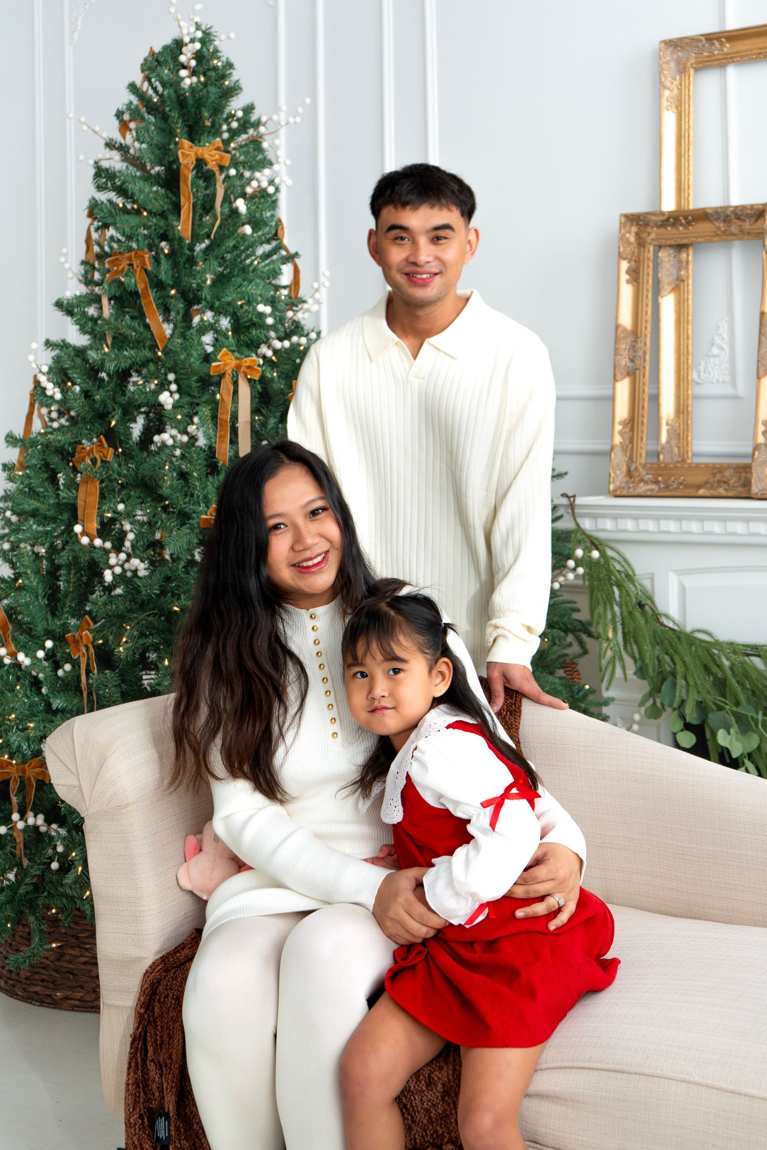 Family of three poses by decorated Christmas tree, parents in white sweaters with daughter in red dress.