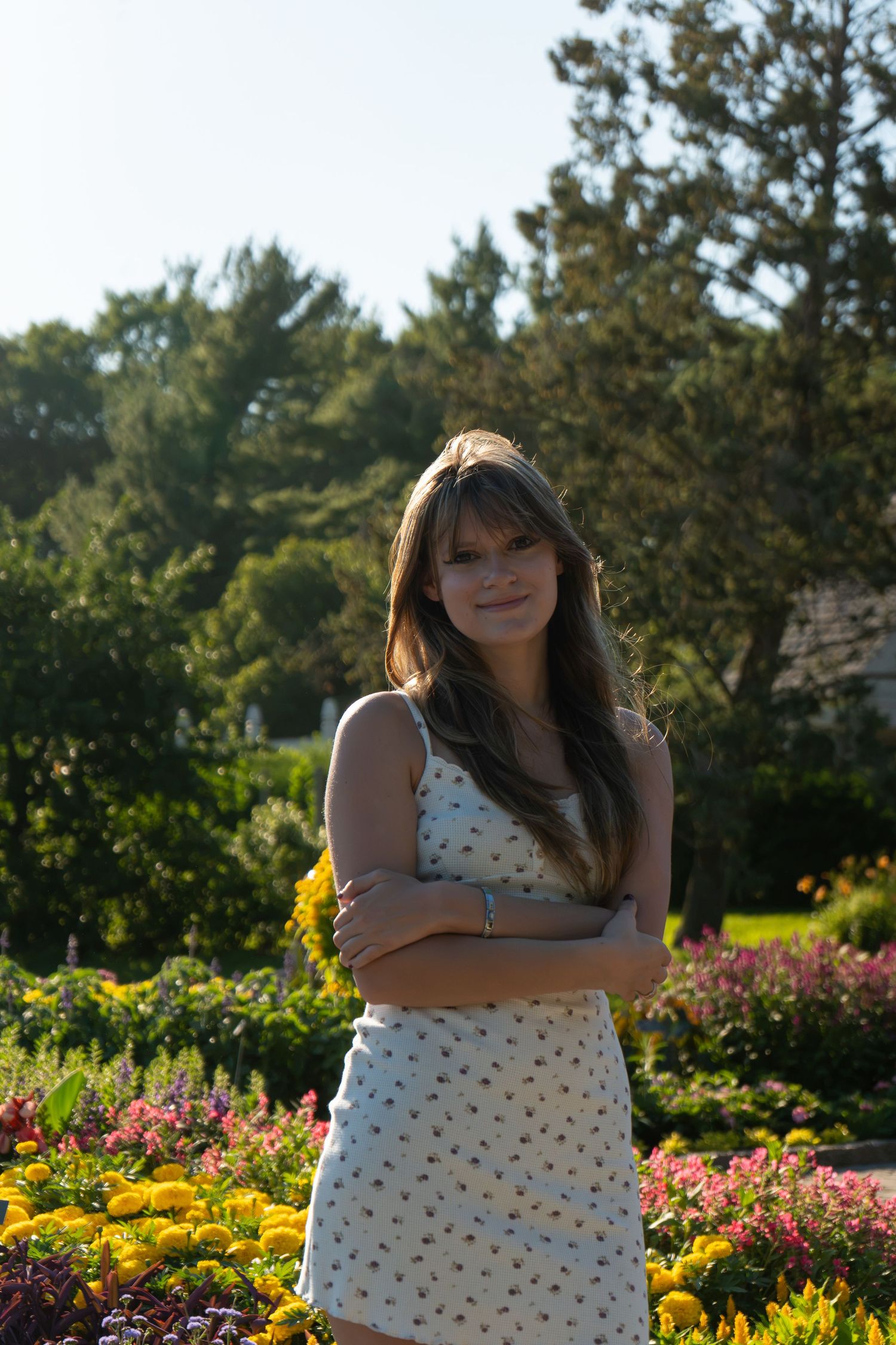 Young woman in floral sundress standing with arms crossed in a colorful garden with blooming flowers and trees.