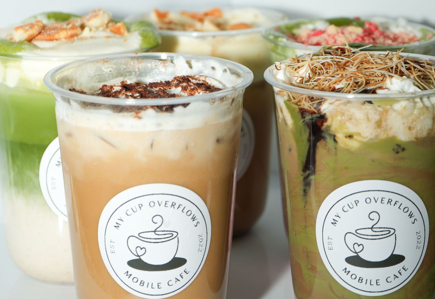 Assorted drinks from My Cup Overflows Mobile Cafe, featuring iced lattes and matcha bowls in branded plastic cups.