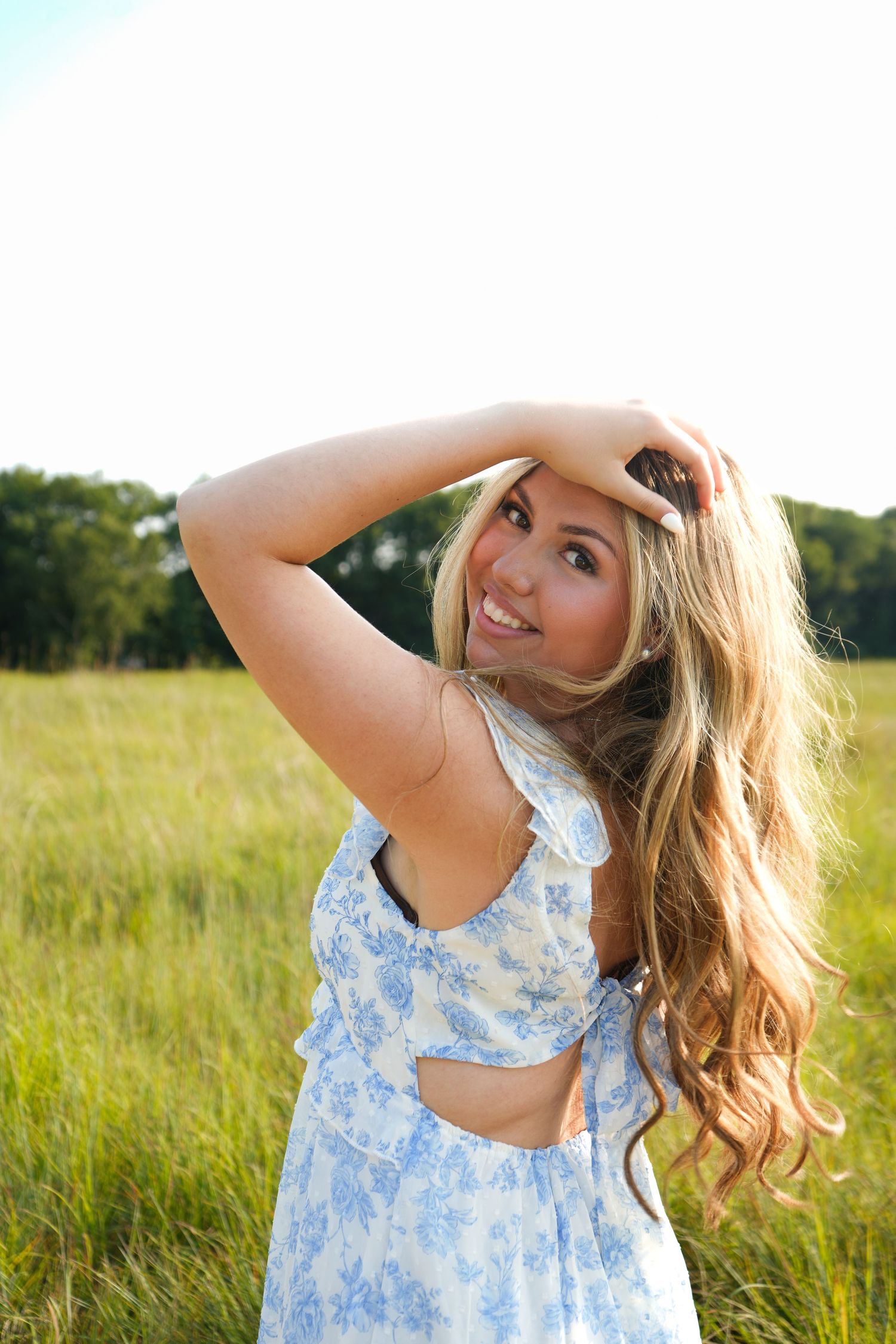 Young blonde woman in blue floral sundress smiling in a sunny green meadow with trees in the background.
