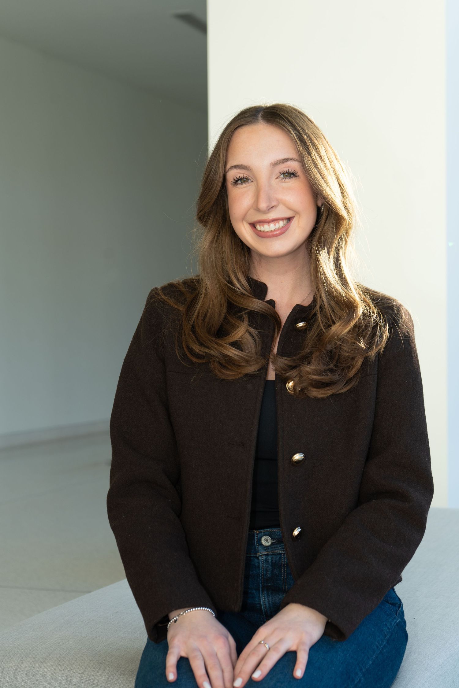 Smiling young woman with long brown hair wearing a dark blazer and jeans, seated in a bright modern space.