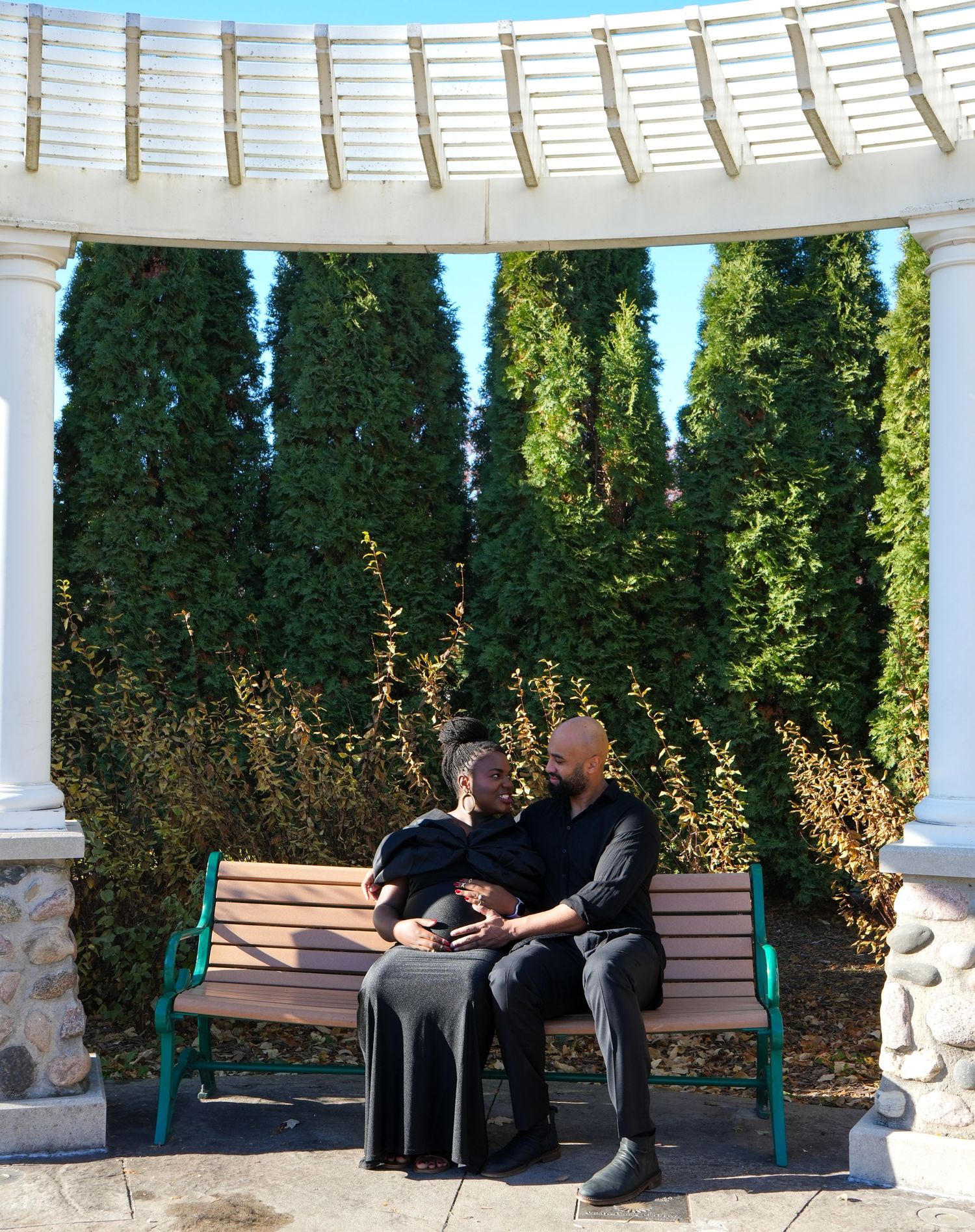 Couple sitting together on a park bench under a white pergola surrounded by tall green cypress trees.