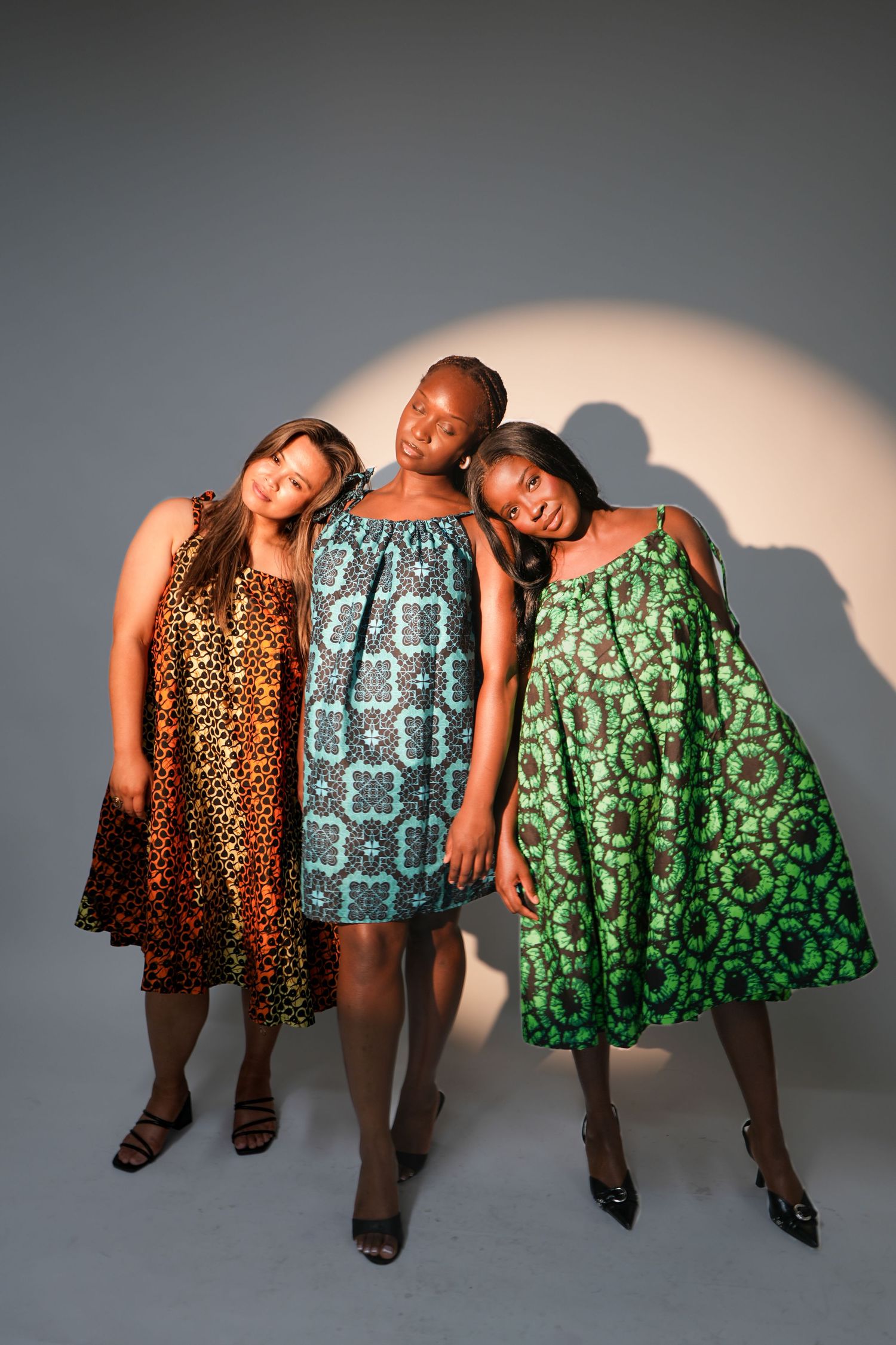 Three women in vibrant African print dresses pose together in a studio, showcasing colorful fashion designs.