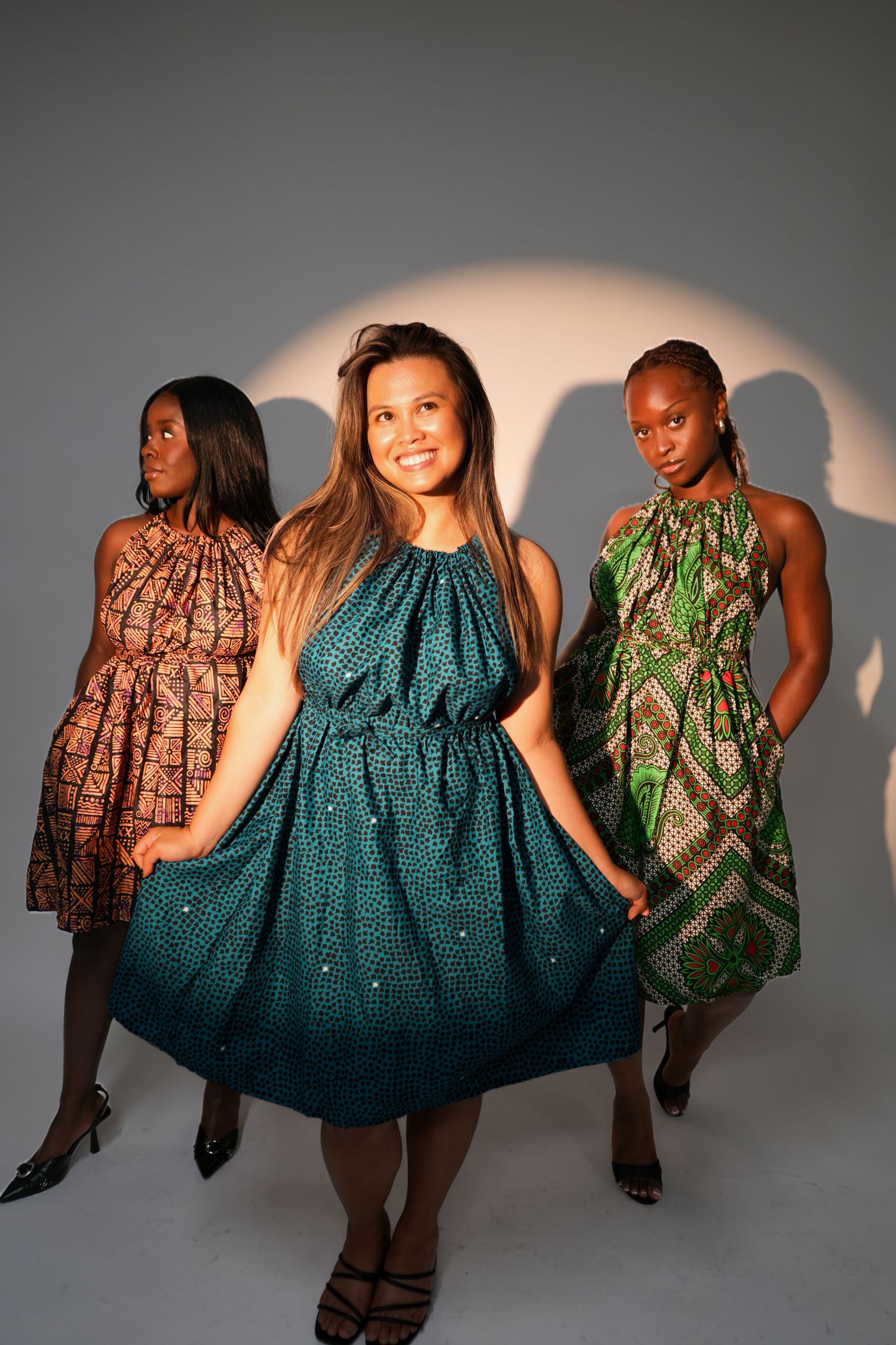 Three women in colorful African print halter dresses pose confidently in a studio with dramatic lighting.