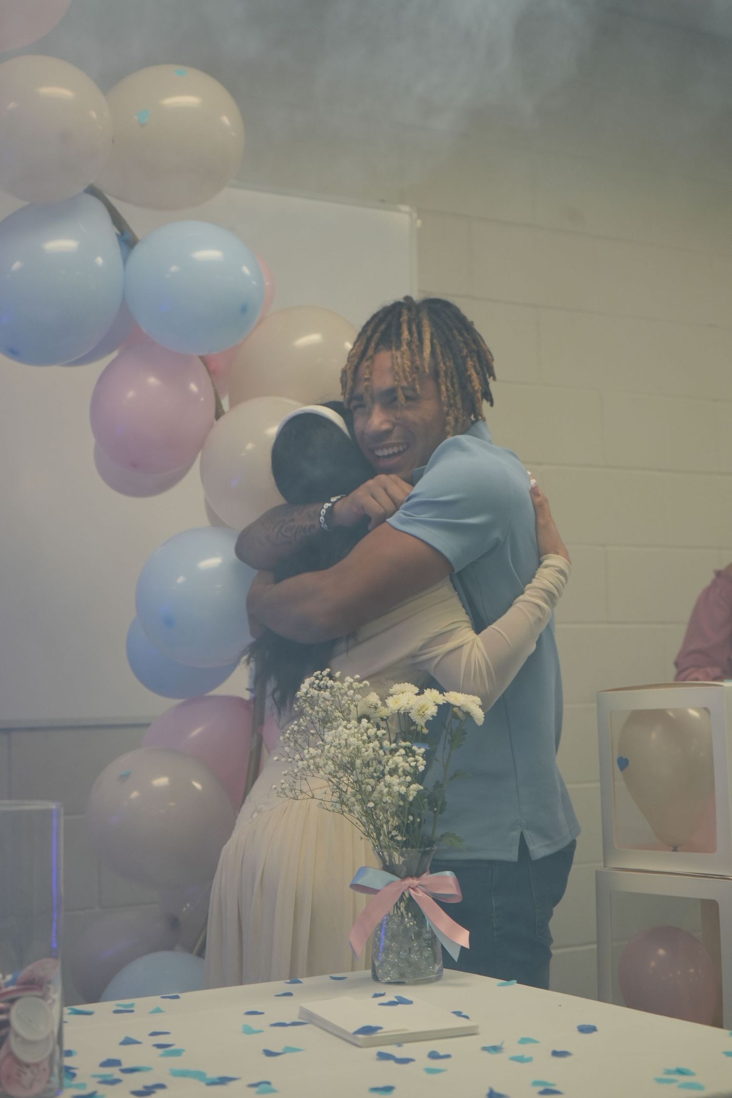 Couple embracing at a gender reveal party with pink and blue balloons and confetti on the table.