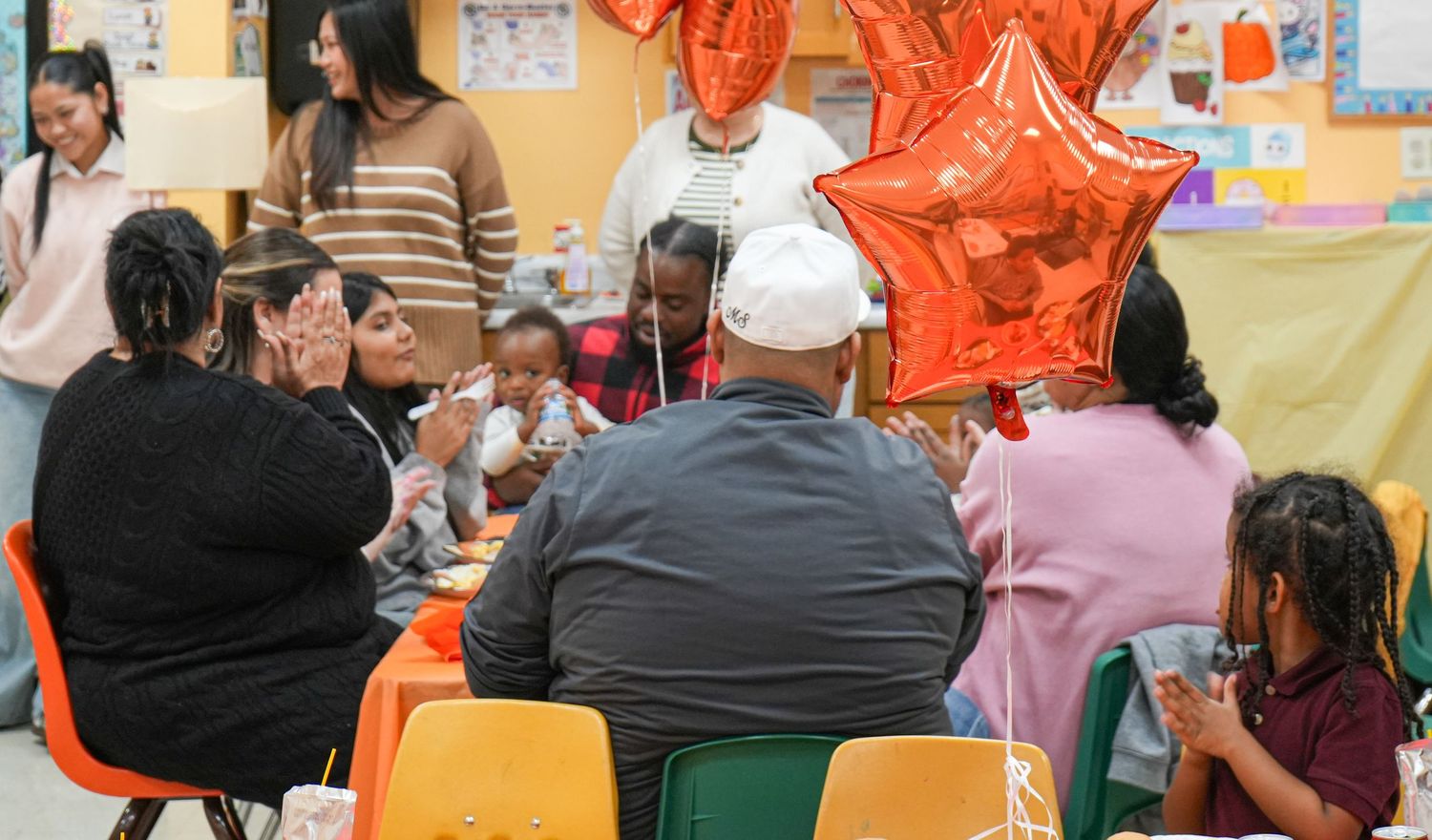 Group of people celebrating with orange star balloons at a colorful indoor party gathering around a table.