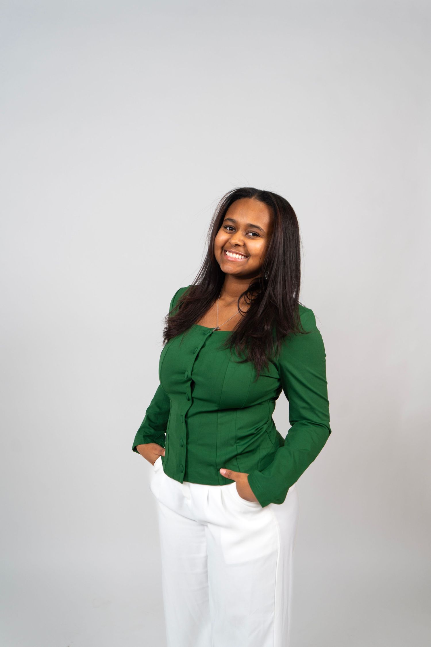 Smiling young woman in green jacket and white pants posing confidently against a light gray background.