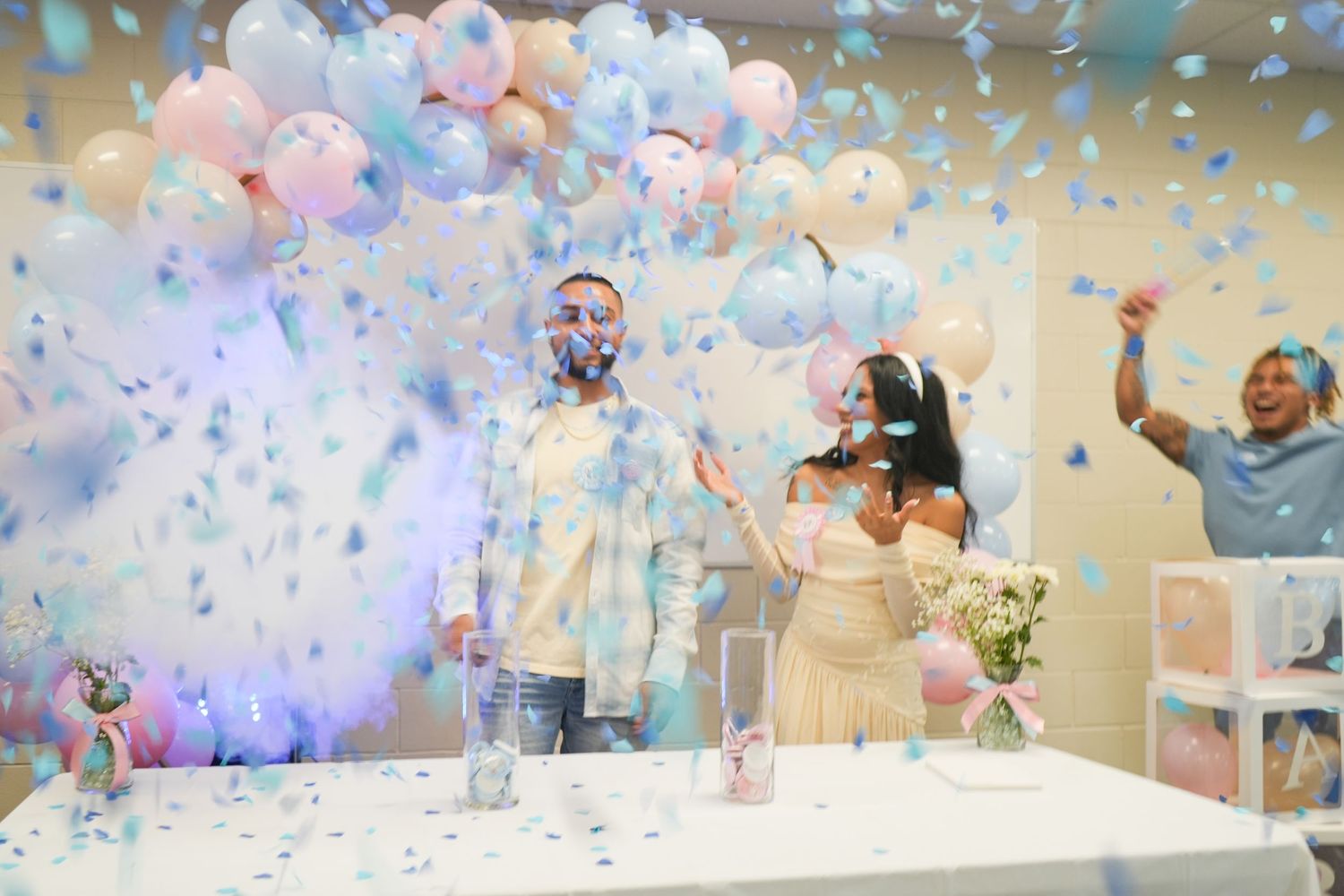 Couple celebrating a gender reveal party with blue confetti, balloons, and guests cheering in a decorated room.