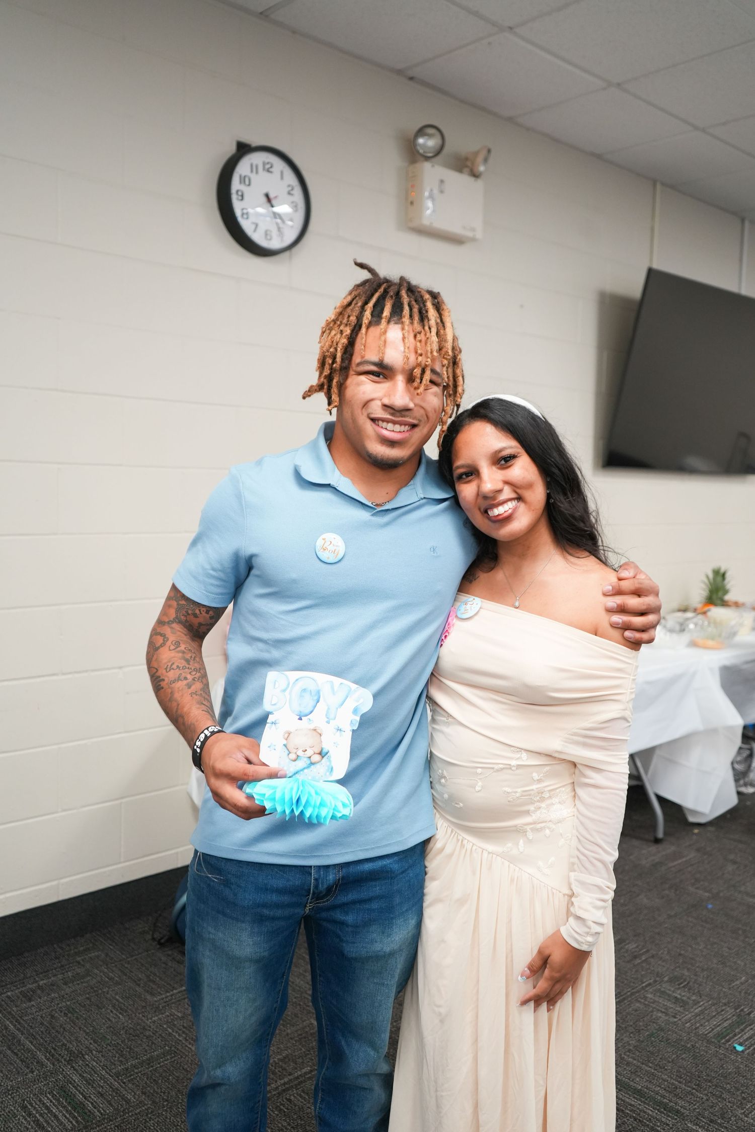 Couple posing together at a baby shower, man in blue polo with tattoos, woman in white off-shoulder dress.