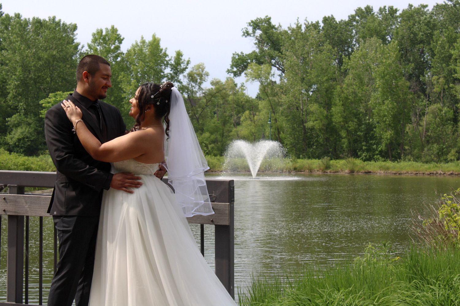Bride and groom embrace on a wooden deck overlooking a serene pond with a fountain and lush green trees.