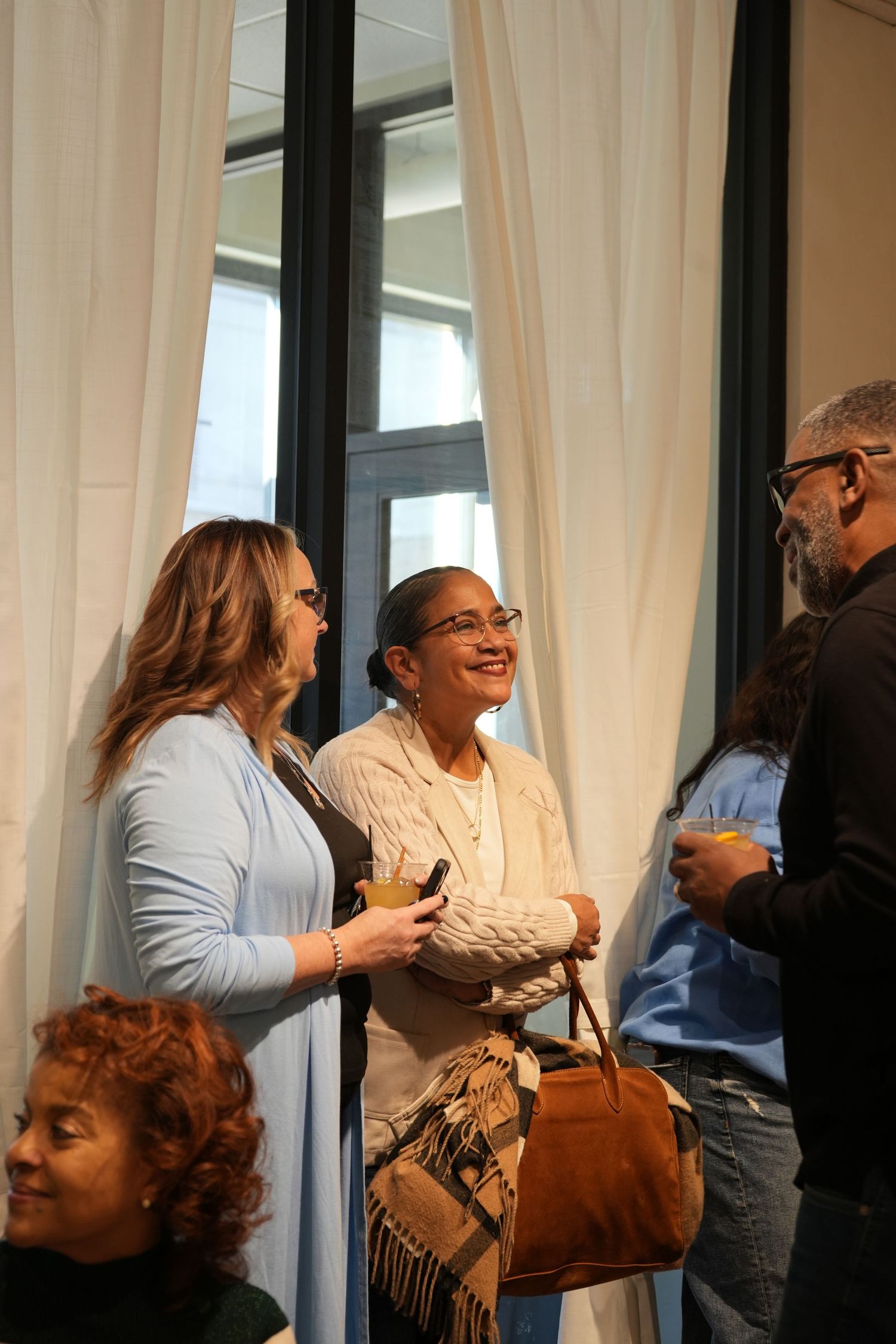 Women smiling and socializing at an indoor gathering near large windows with white curtains.