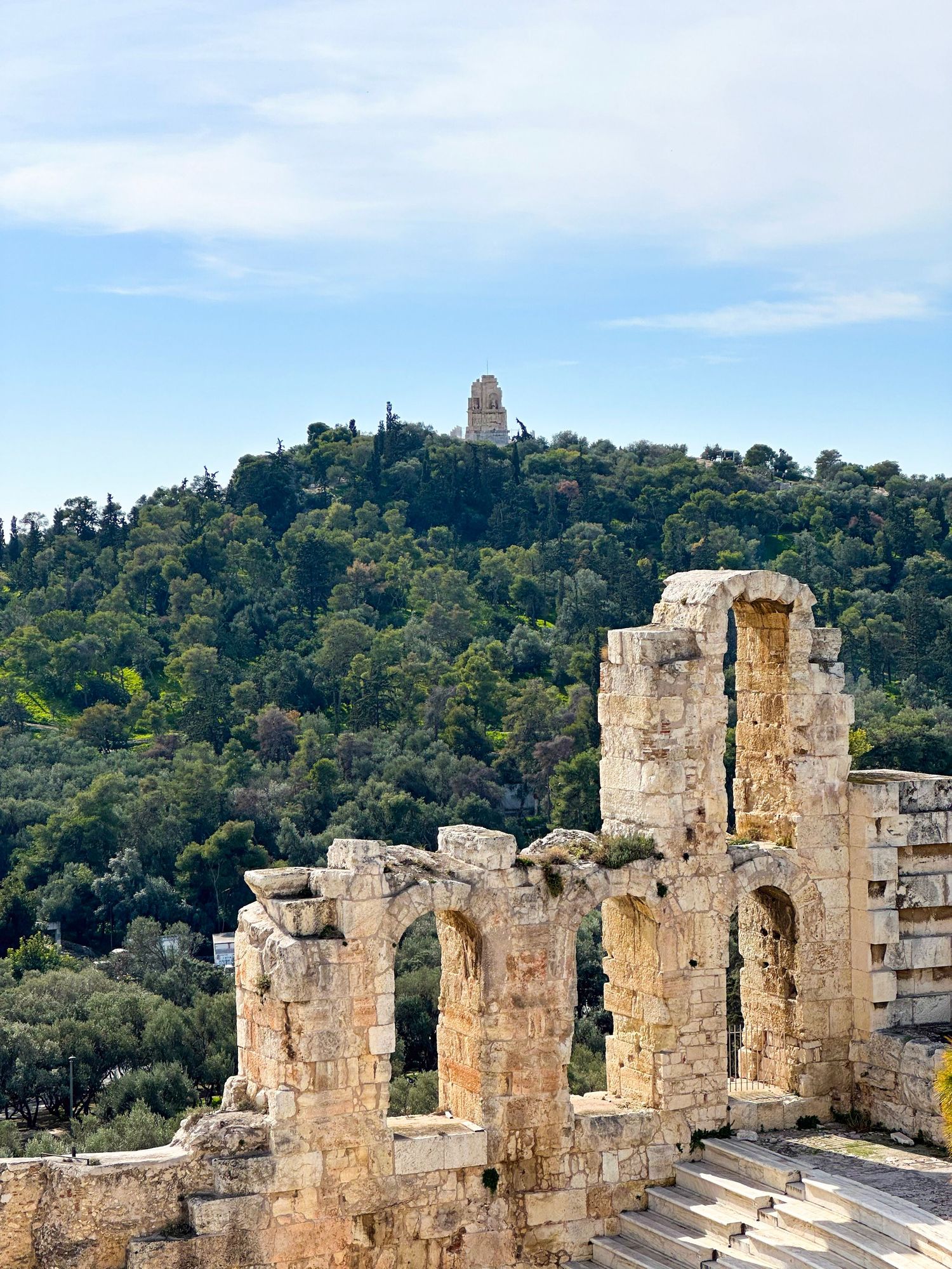 Ancient Greek ruins with stone arches on the Acropolis, overlooking a lush green hill with a monument in Athens, Greece.