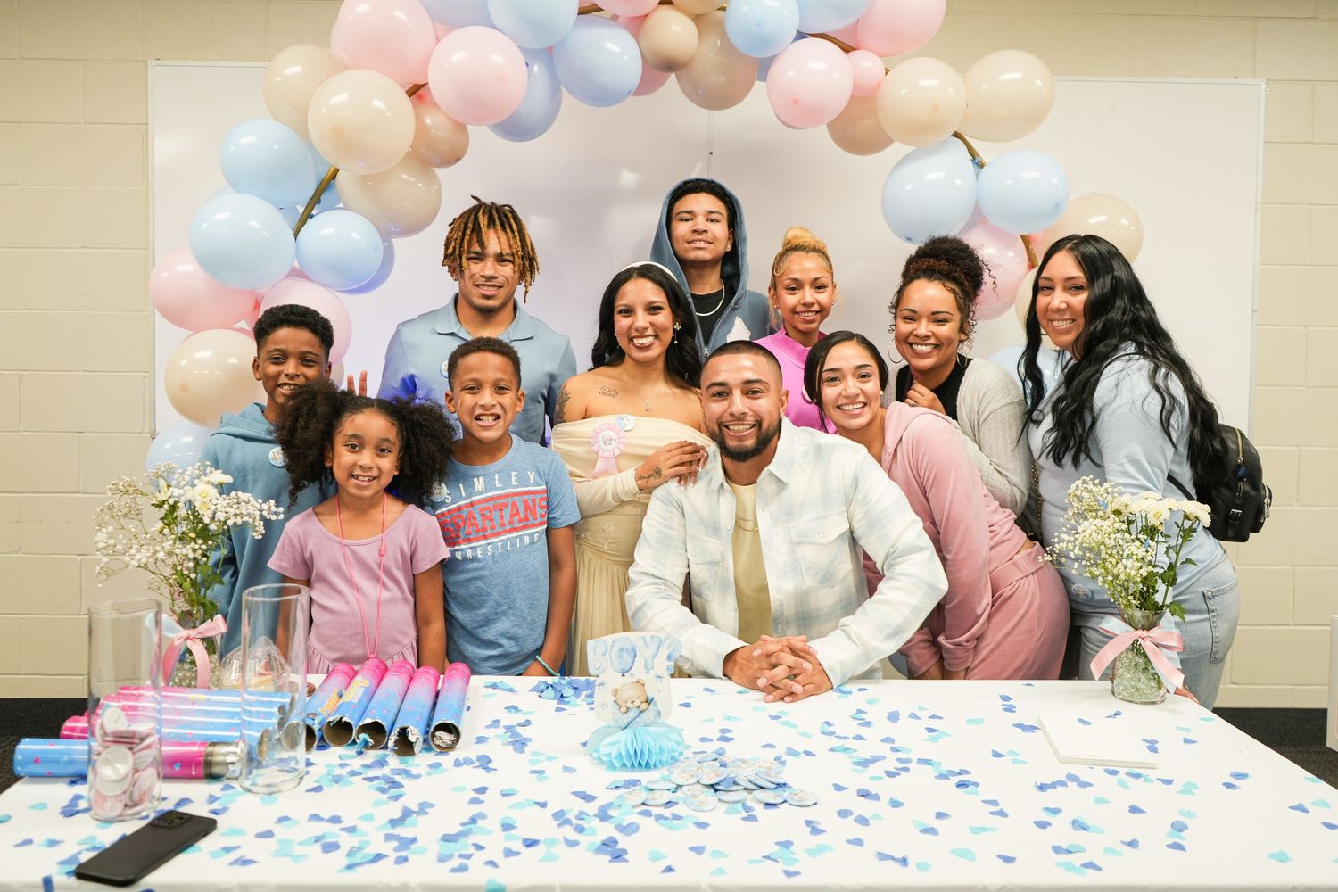 Group of family and friends celebrating a gender reveal party with blue confetti and pink and blue balloon arch.