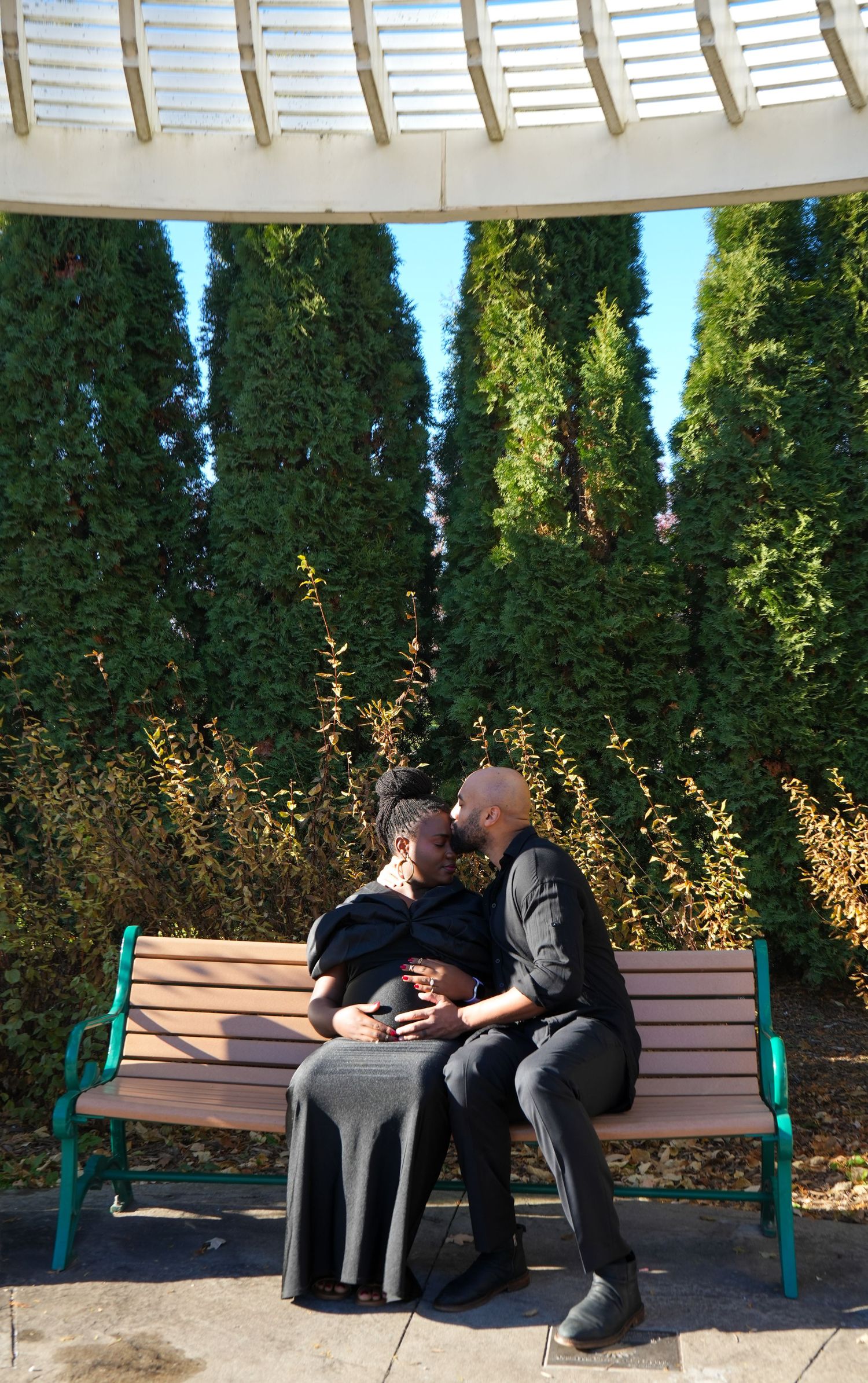 Couple sitting close together on a wooden bench in a garden gazebo, surrounded by tall green trees on a sunny day.
