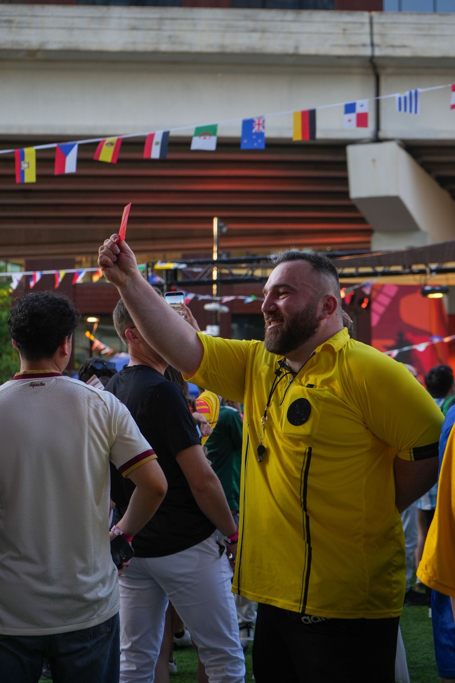 Man in yellow referee shirt holds red card aloft at outdoor sports viewing event with international flags.