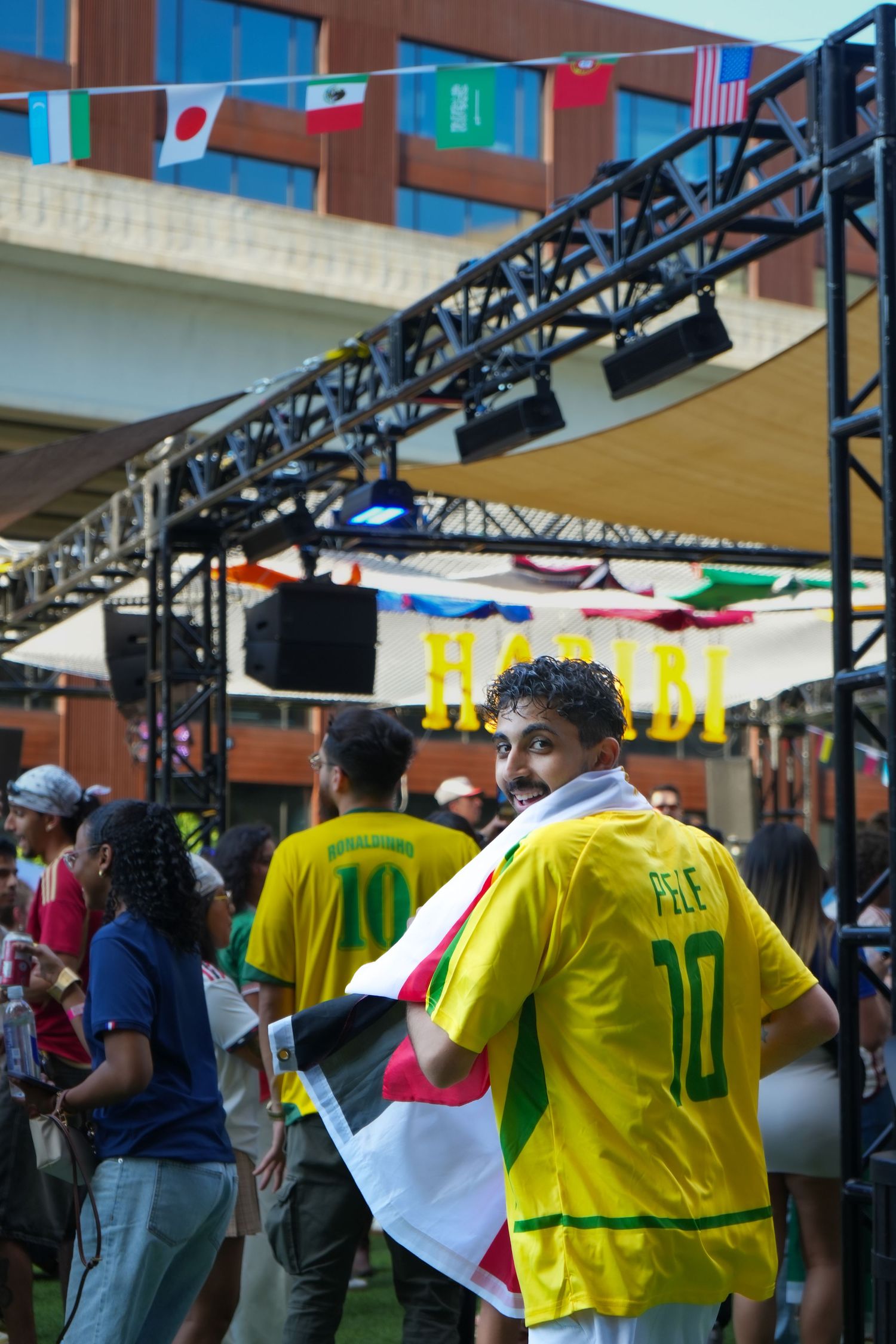 Soccer fan wearing Brazil jersey number 10 with flag draped over shoulders at outdoor World Cup viewing party.