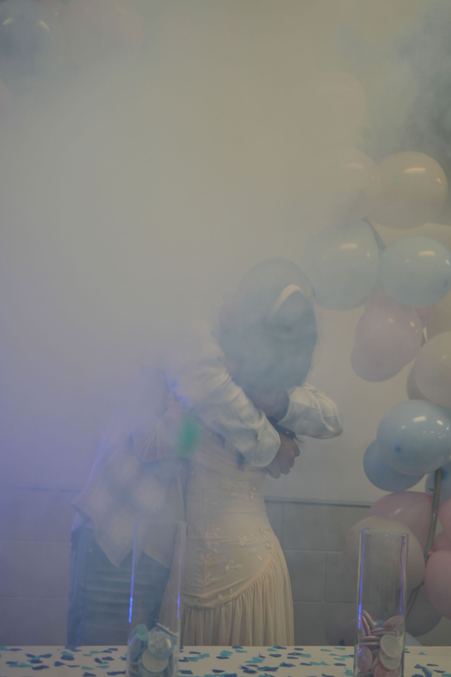 Gender reveal party with blue smoke, balloons, and confetti surrounding a couple in a festive decorated venue.