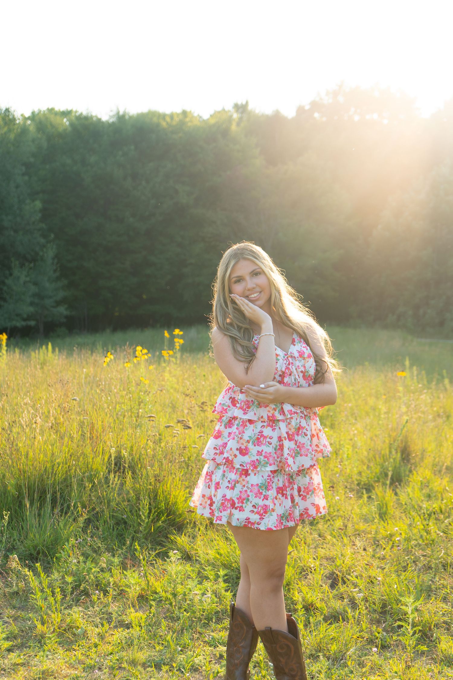 Young blonde woman in floral dress and cowboy boots posing in sunlit meadow with wildflowers and trees.