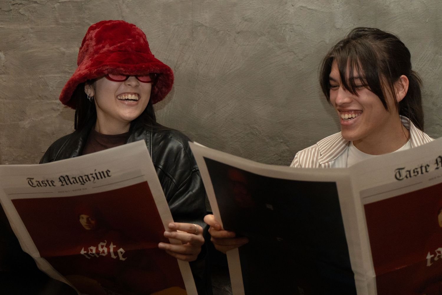 Two women smiling and reading Taste Magazine together against a gray wall.