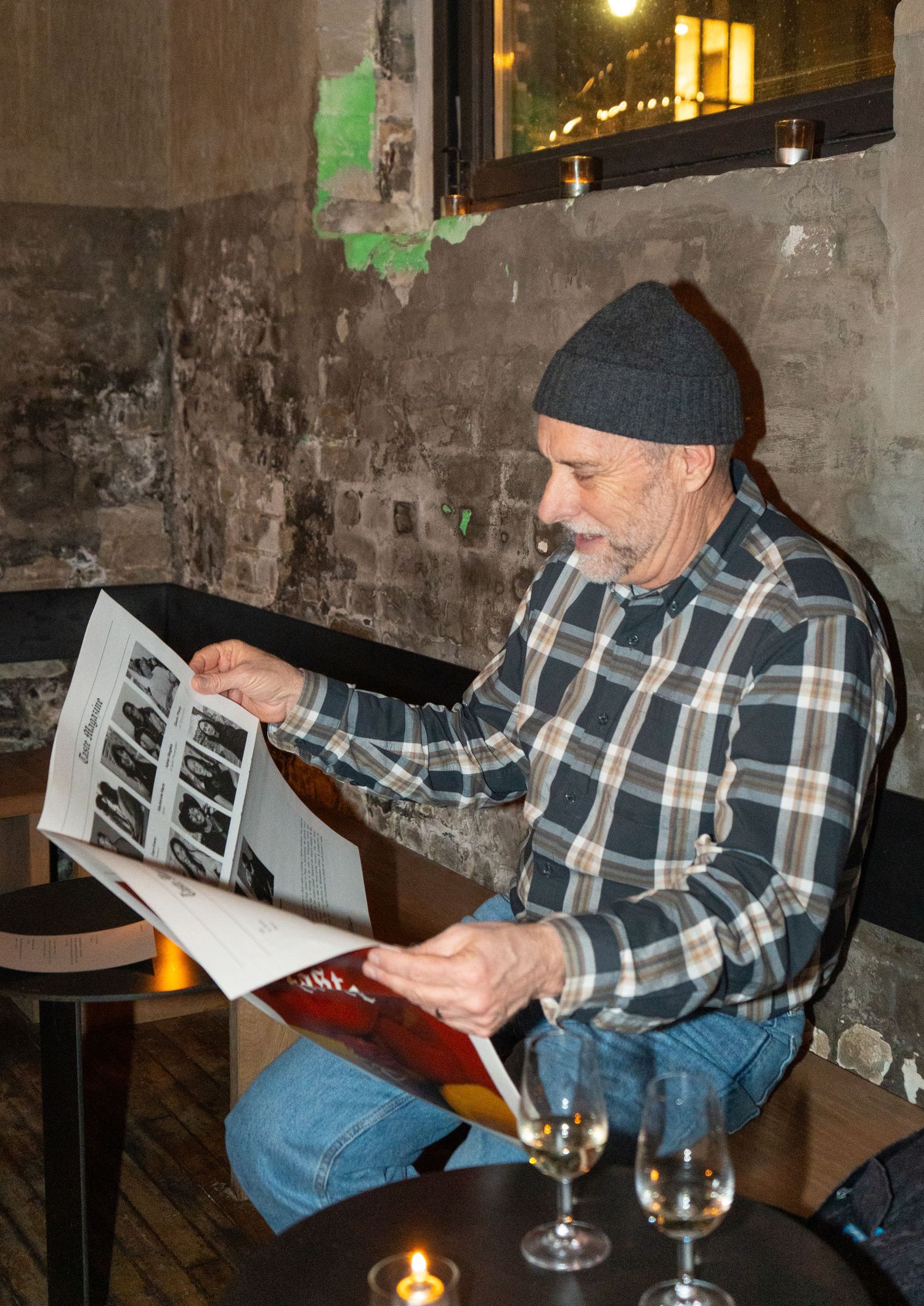 Man in plaid shirt and beanie reads a magazine at a rustic bar table with wine glasses and candle.